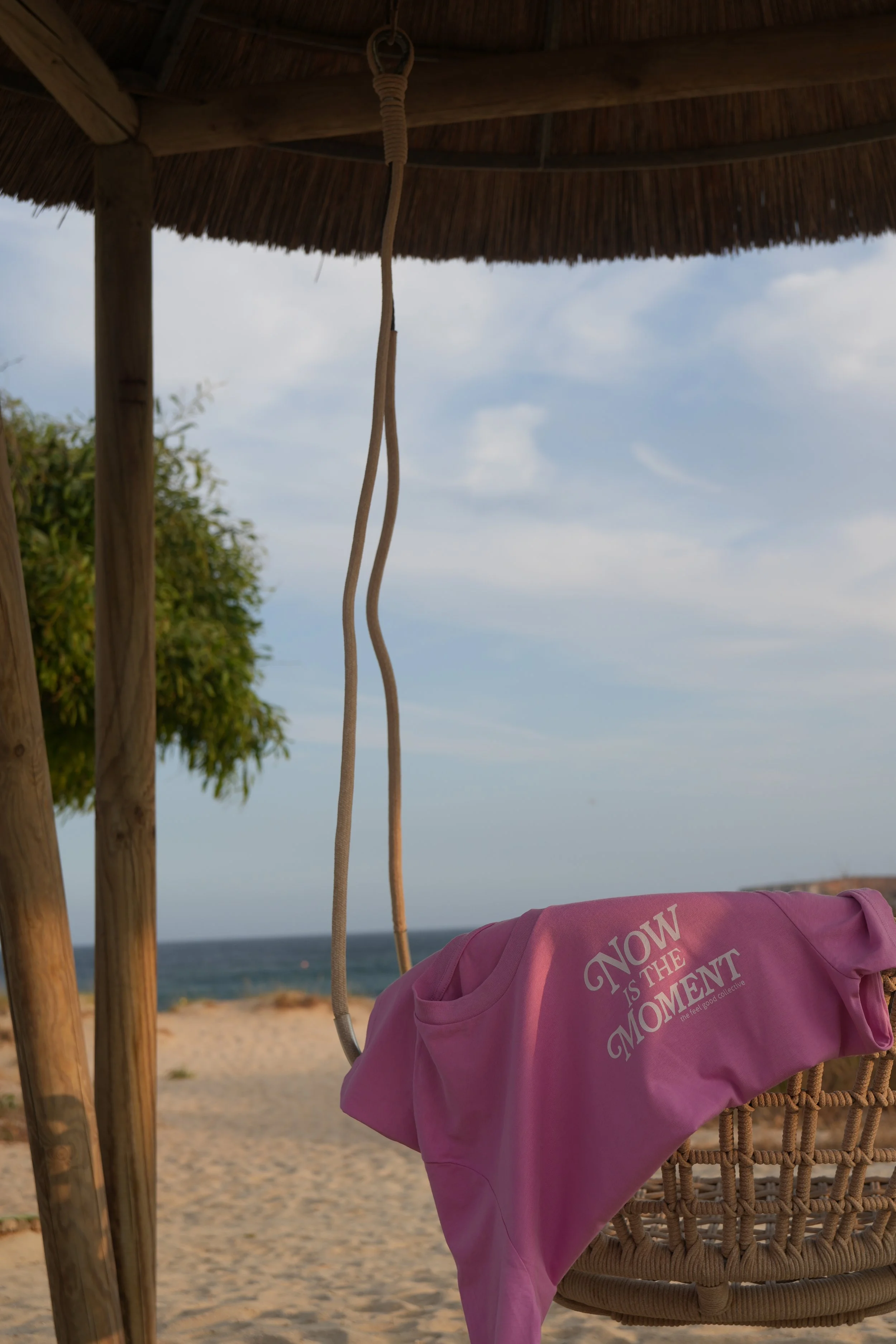 Blick auf einen Strand mit einem hängenden Korbsessel unter einem Strand- oder Garten-Pavillon mit einem pinken T-Shirt darauf, das „NOW IS THE MOMENT“ steht, und das Meer im Hintergrund bei bewölktem Himmel.