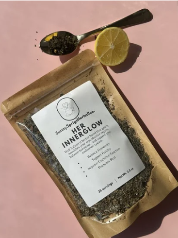 A bag of Sunny Springs Herb Tea Inner Glow with a lemon half, a lemon slice, and a spoon with tea leaves on a pink background.