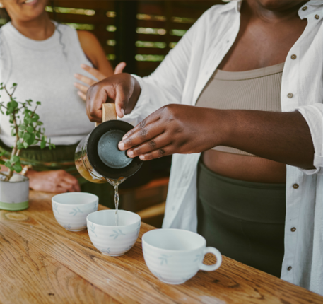 Person pouring tea into cups on a wooden table with another person in the background.