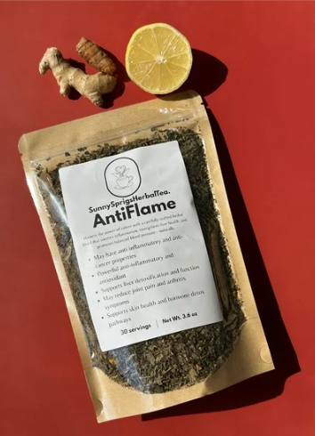 A package of SunnySpringsHerbalTea AntiFlame herbal tea on a red surface, with a lemon half and a piece of ginger root beside it.