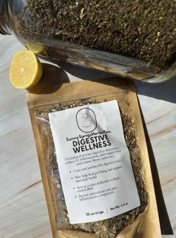 A packet of Sunny Sprout Herbal Tea labeled 'Digestive Wellness' placed on a light-colored surface, with half a lemon and a container of soil or herbs nearby.