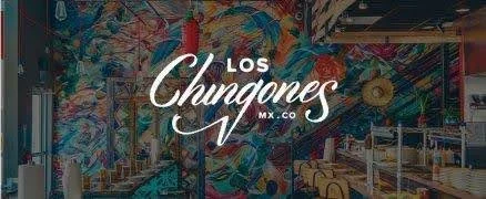 Interior of a colorful cafe or restaurant with vibrant wall art, tables, chairs, and a neon sign that reads 'Los Chingones'MX.CO'.