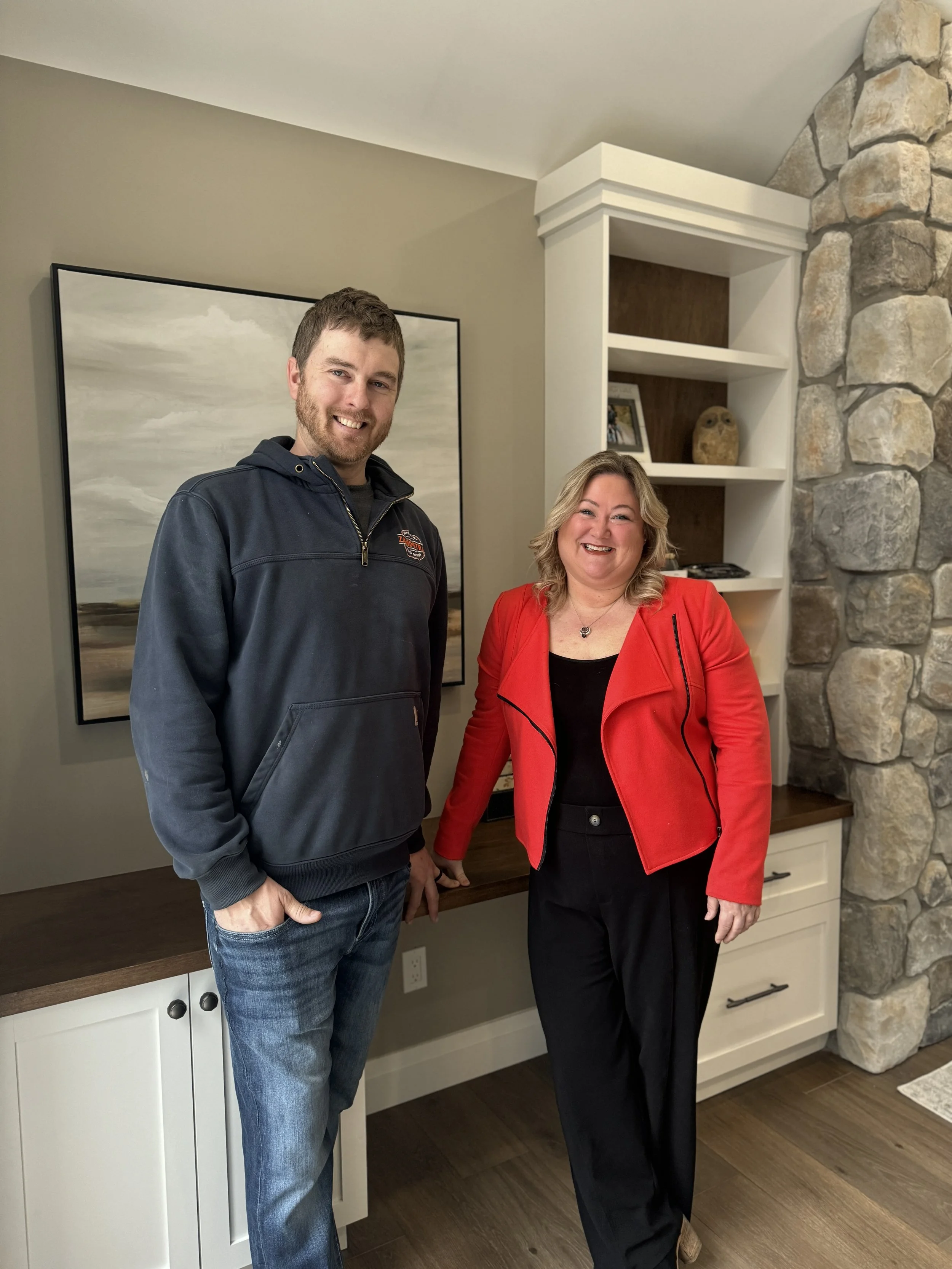 A man and a woman are standing together in a living room, smiling at the camera. The man is wearing a black hoodie and jeans, the woman is wearing a red jacket and black pants.