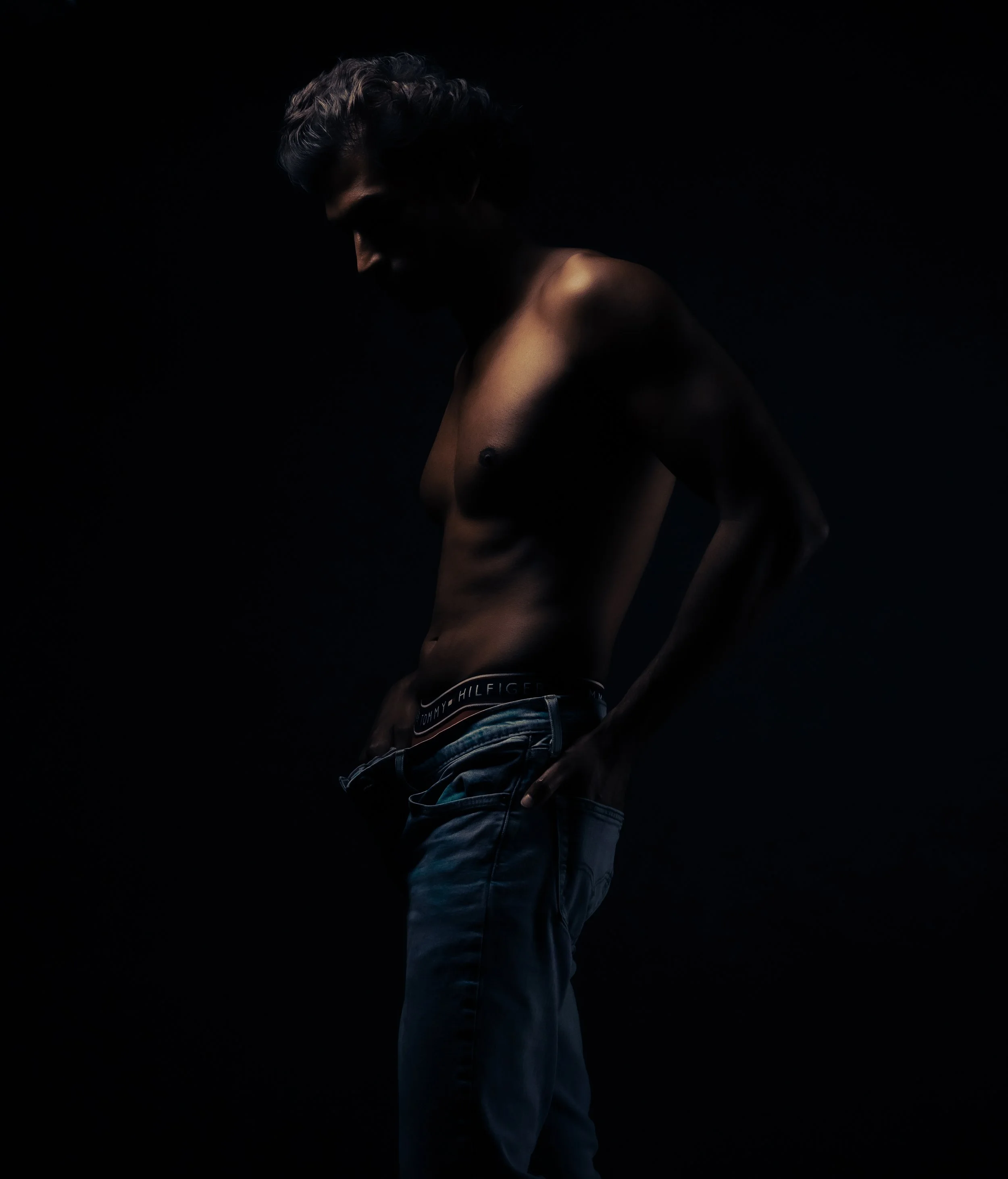A shirtless man with dark hair and a beard, wearing jeans with a Tommy Hilfiger waistband, standing against a black background with dramatic lighting highlighting his upper body and face in profile.