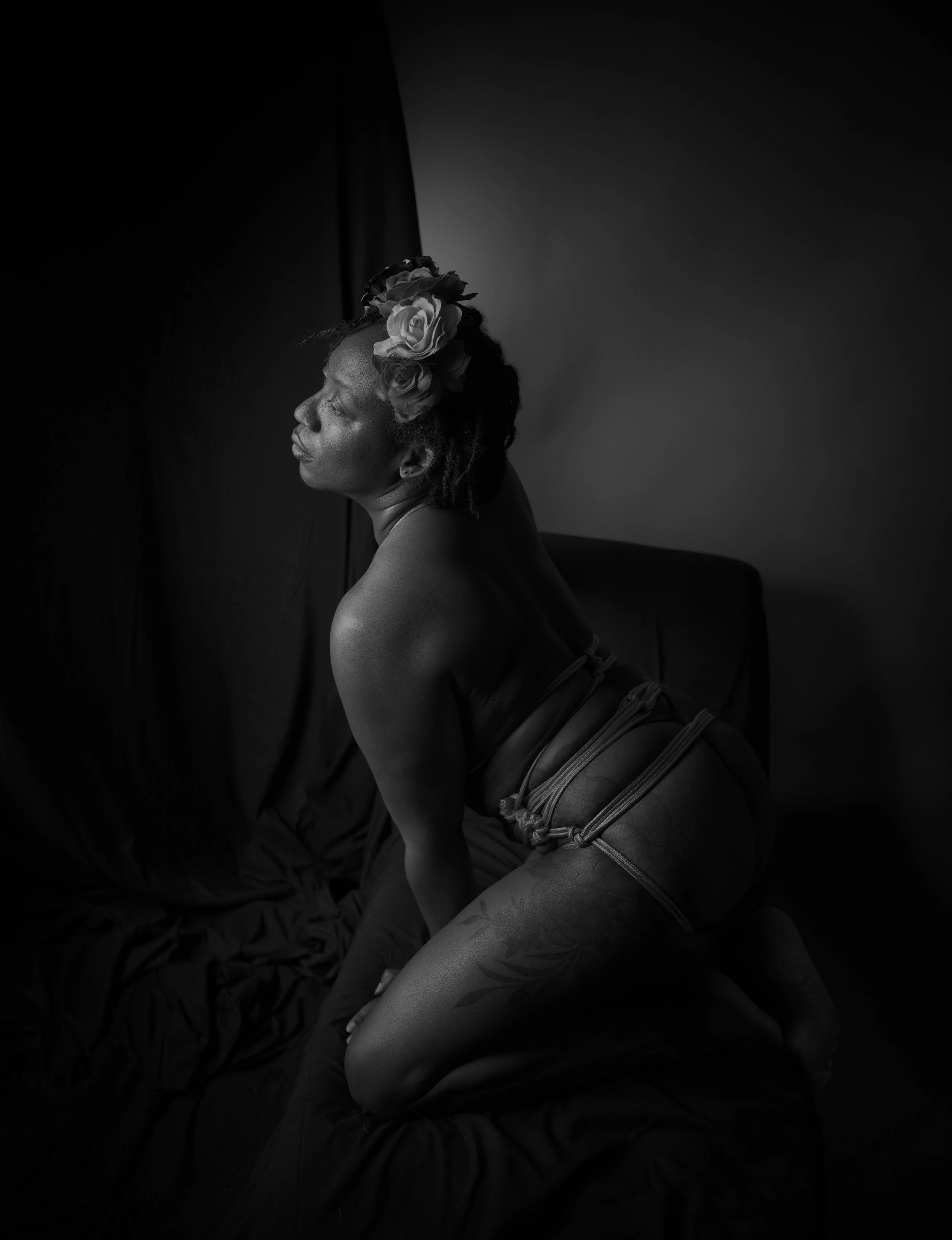 Black and white photo of a woman with flowers in her hair, kneeling on a bed with dark sheets, looking to the side with eyes closed.