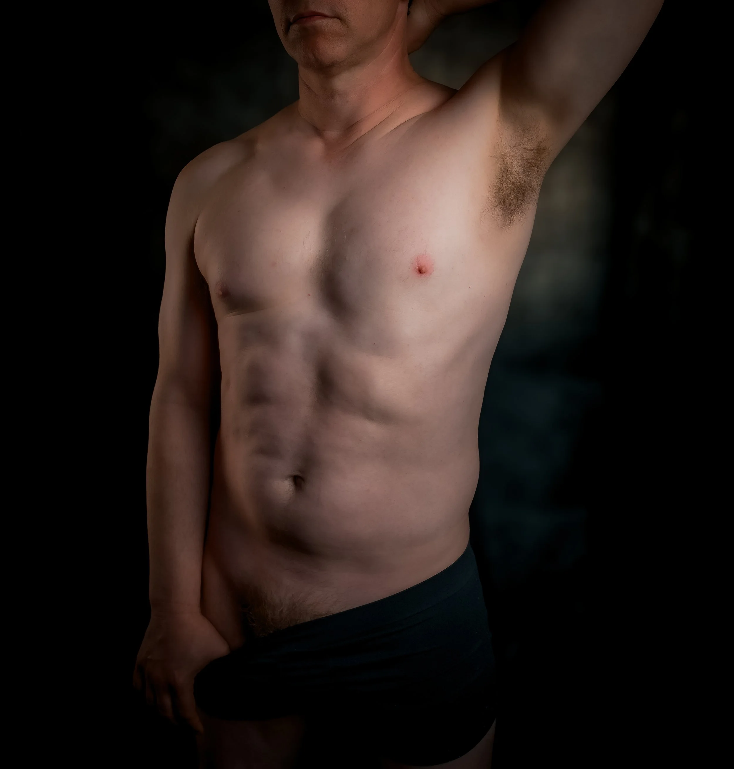A shirtless man with one arm raised behind his head, standing against a dark background.