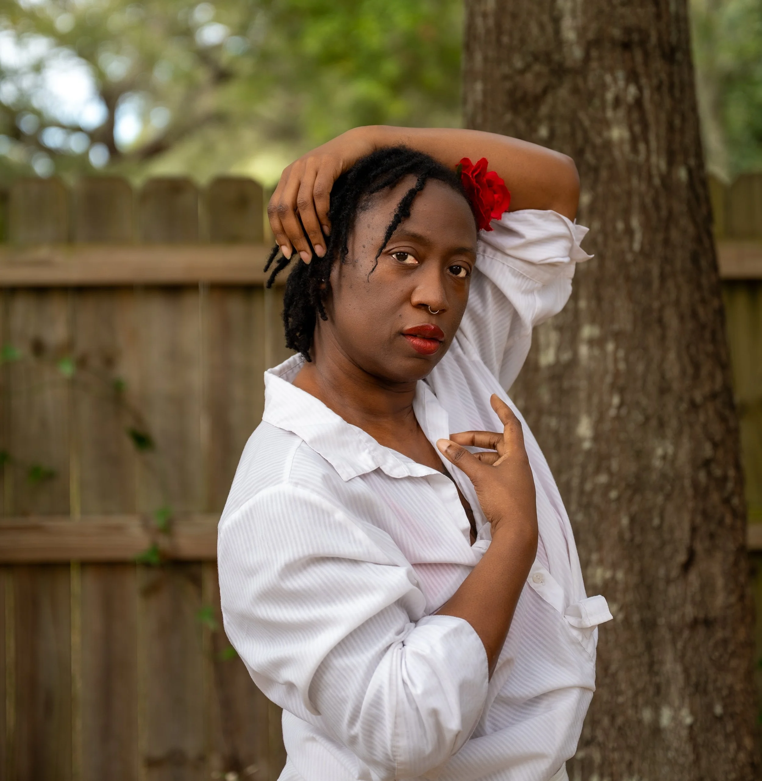 A woman with dark skin and dreadlocks, wearing a pink flower headband, purple dress, and black shiny thigh-high boots, sitting on a beige chaise lounge with a cream satin backdrop.