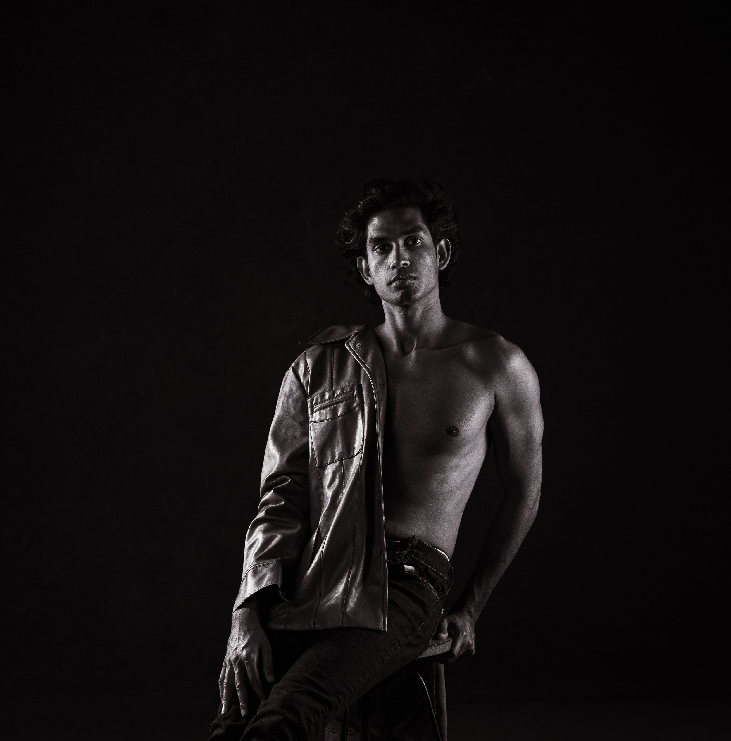 Black and white studio portrait of a shirtless young man with a leather jacket draped over one shoulder, gazing confidently upward against a black background.  Boudoir for Men.  Male Boudoir. Gentlemen Boudoir.  Dudeoir and masculine boudoir.