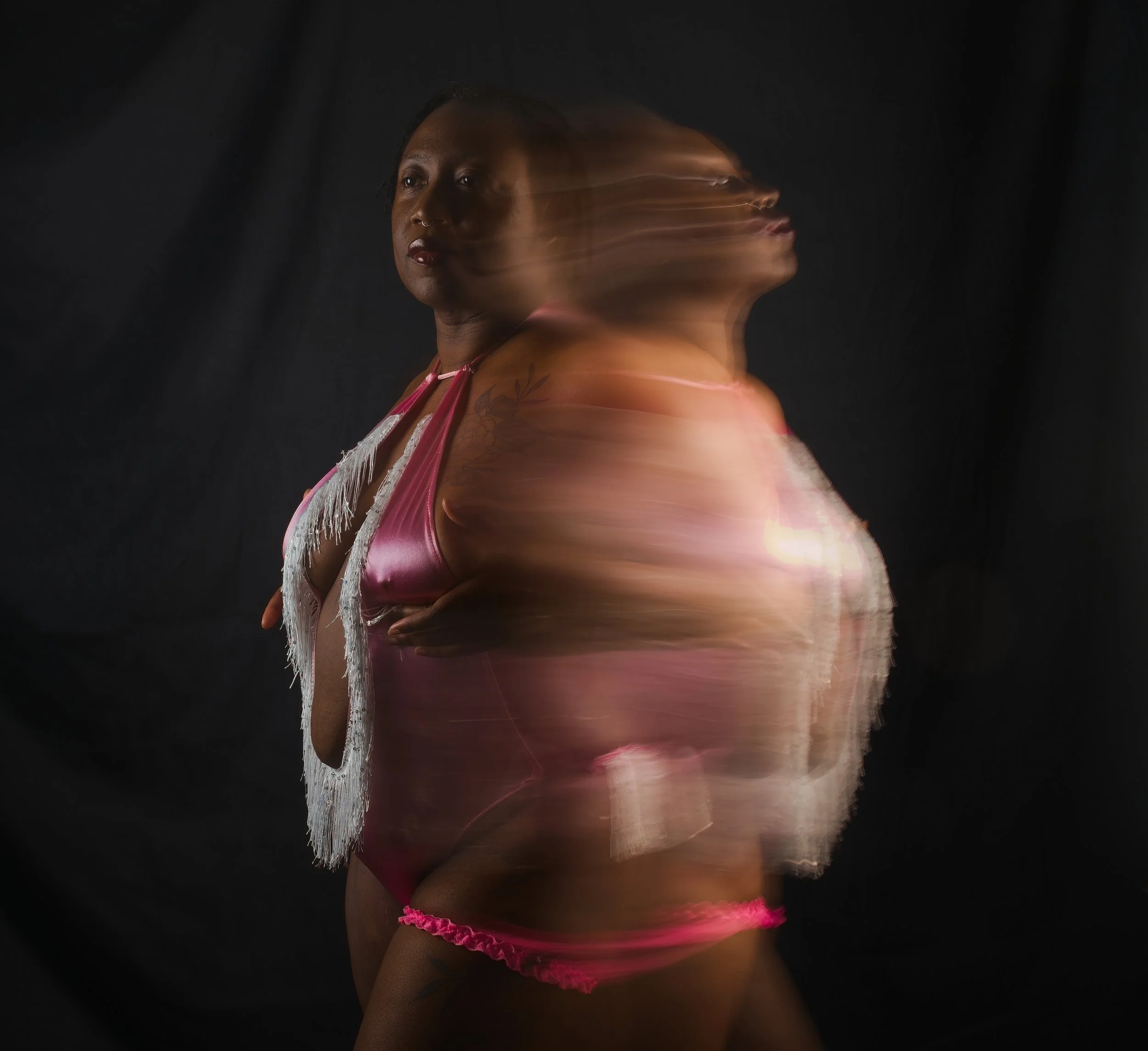 A woman in pink metallic clothing with fringes, standing against a black background, has a motion-blurred twin effect created by long exposure.