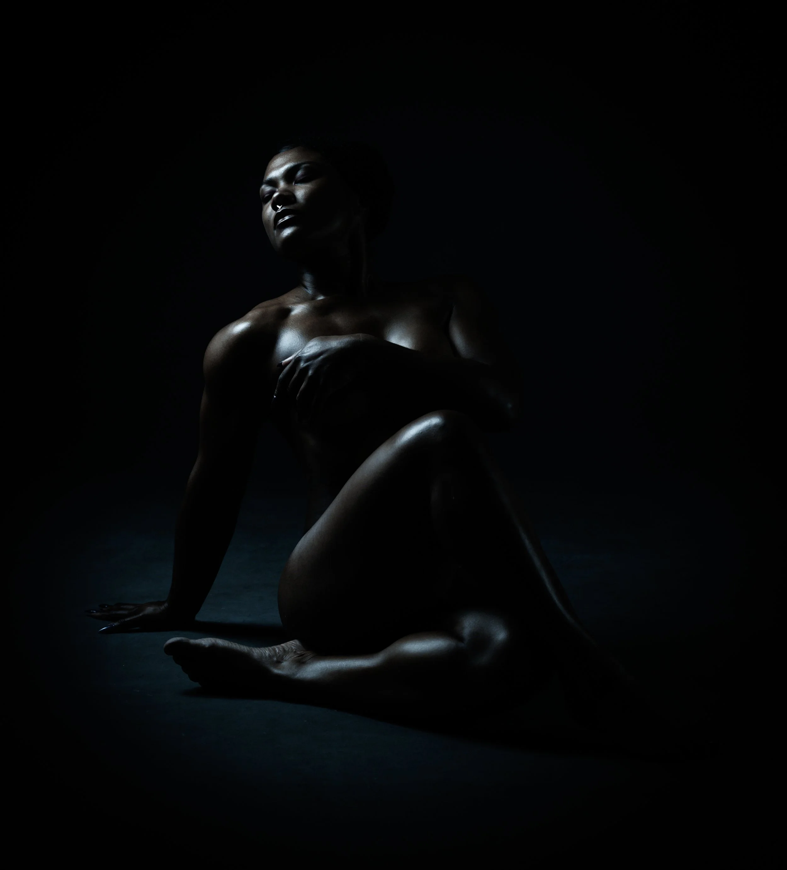 A nude woman with dark skin, short hair, and a septum piercing posed against a dark background, with soft lighting highlighting her face and upper body.