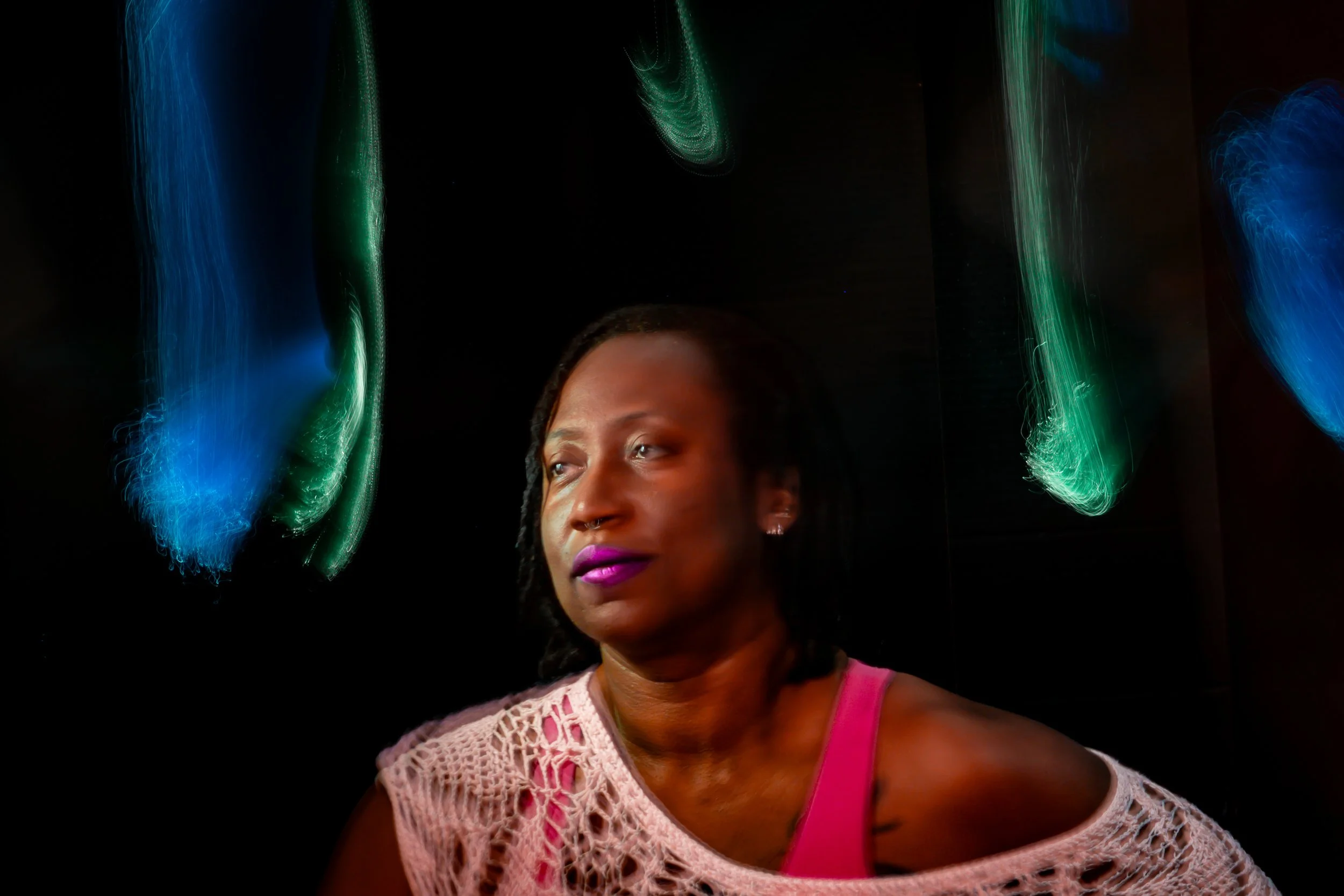 Painterly light painting portrait of a woman looking to the side with purple lipstick, framed by soft blue and green glowing light trails against a black background, wearing a white lace top, Bellissimo Expressions studio.
