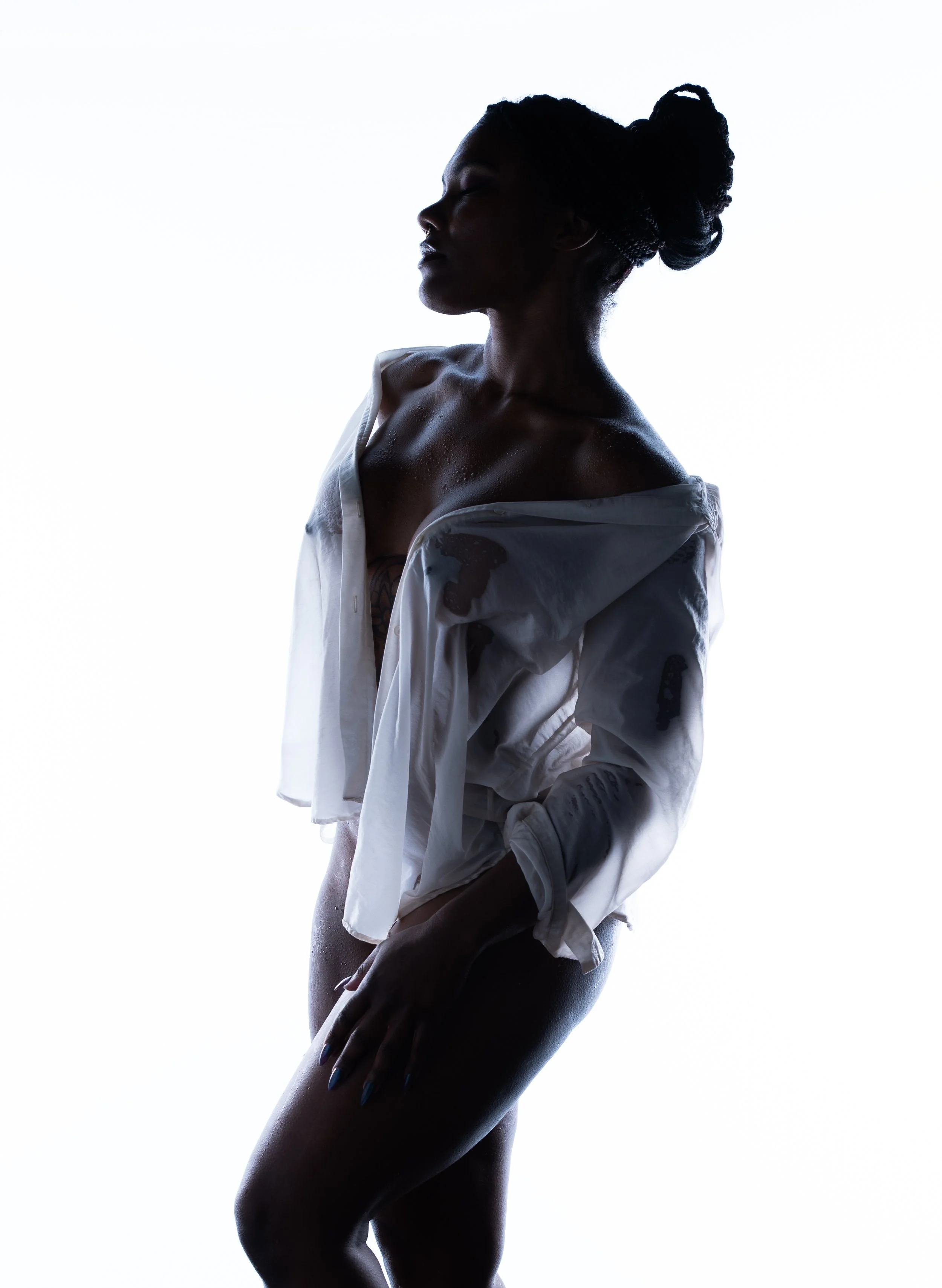 Silhouette of a woman in profile with her eyes closed, wearing an open white shirt, and her hair styled in an intricate updo, with water droplets on her skin against a white background.