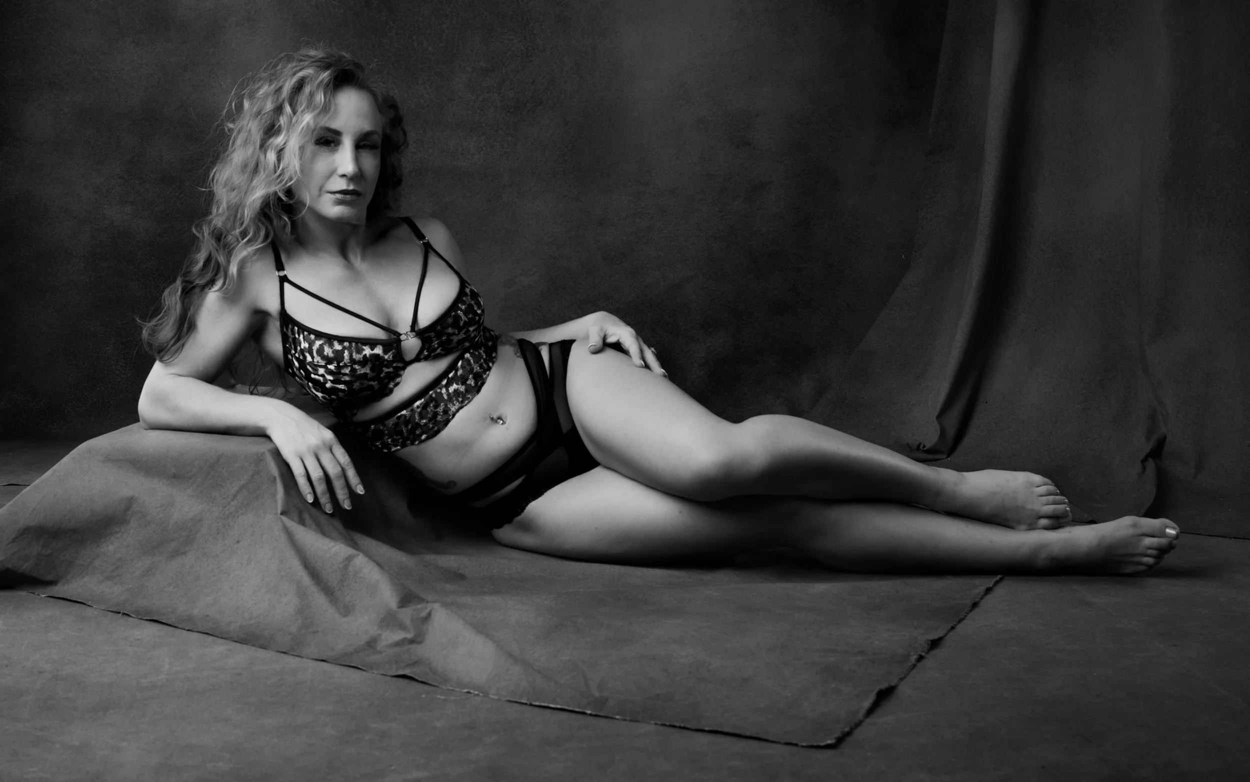 A woman with curly hair lying on a draped surface, wearing lingerie and stockings, in a black and white photoshoot setting.