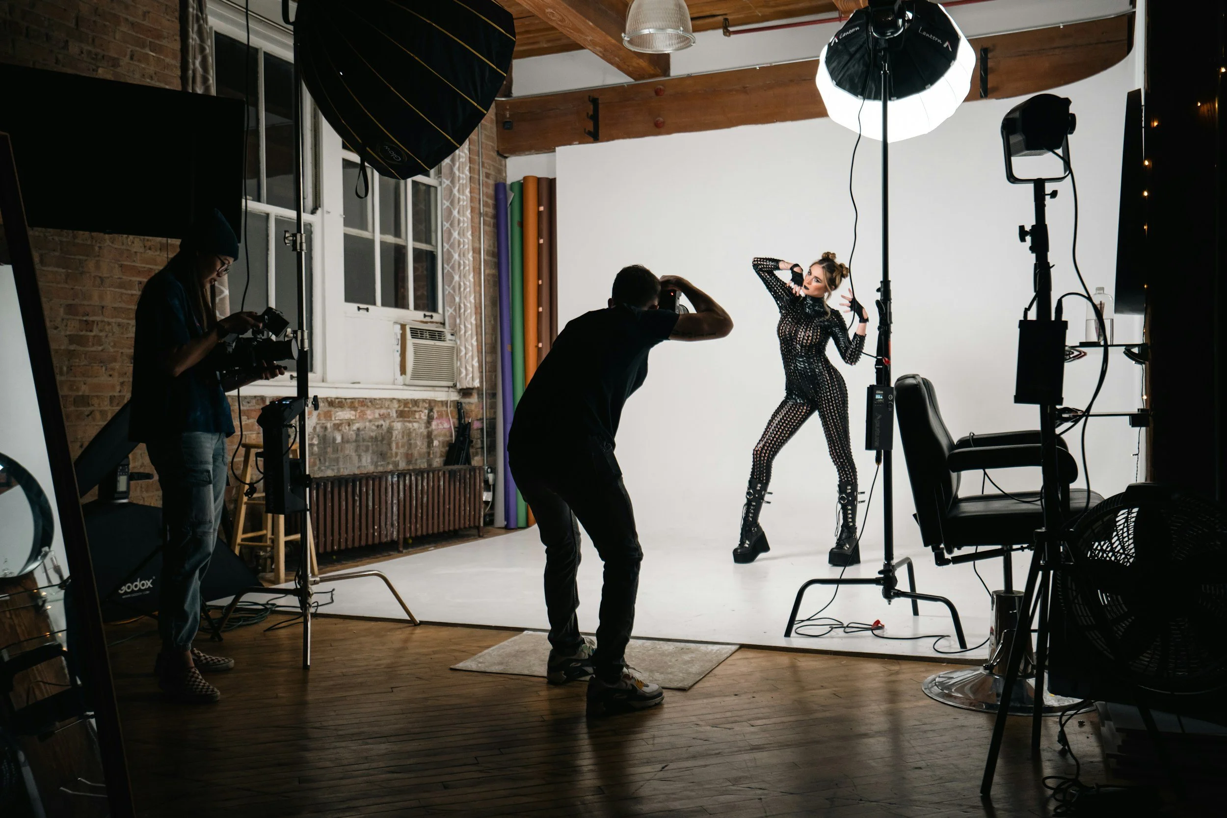 A photoshoot session in a studio with a model in a black, perforated bodysuit and high-heeled boots, posing on a white backdrop. The photographer is taking pictures, and an assistant is managing the lighting equipment. The studio has a wooden ceiling, brick walls, and several lighting and photographic equipment around.