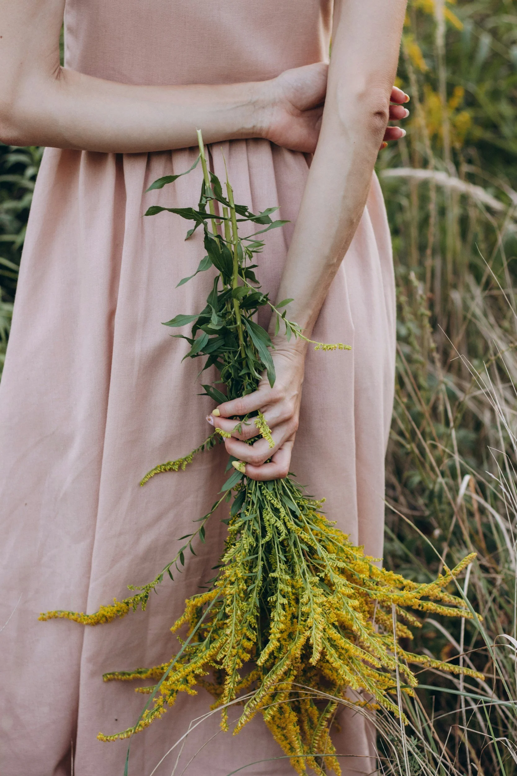 Person in a pink dress holding a bunch of yellow flowering plants in a grassy outdoor setting.