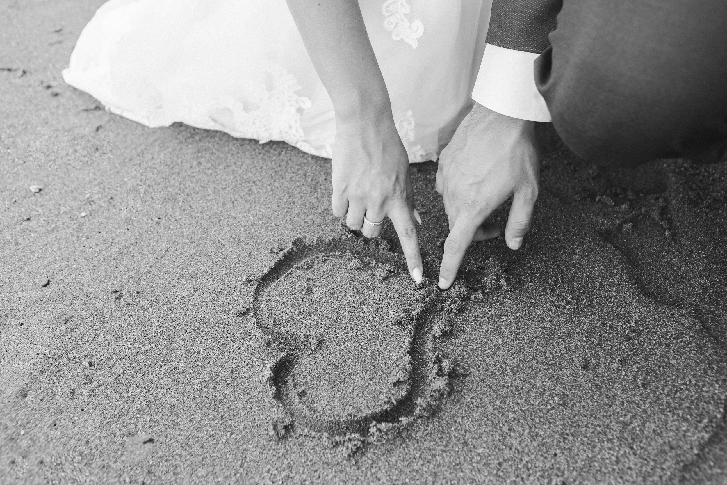 A person wearing a wedding dress and umbrella is drawing a heart in the sand with their finger, next to another person who is pointing at it.