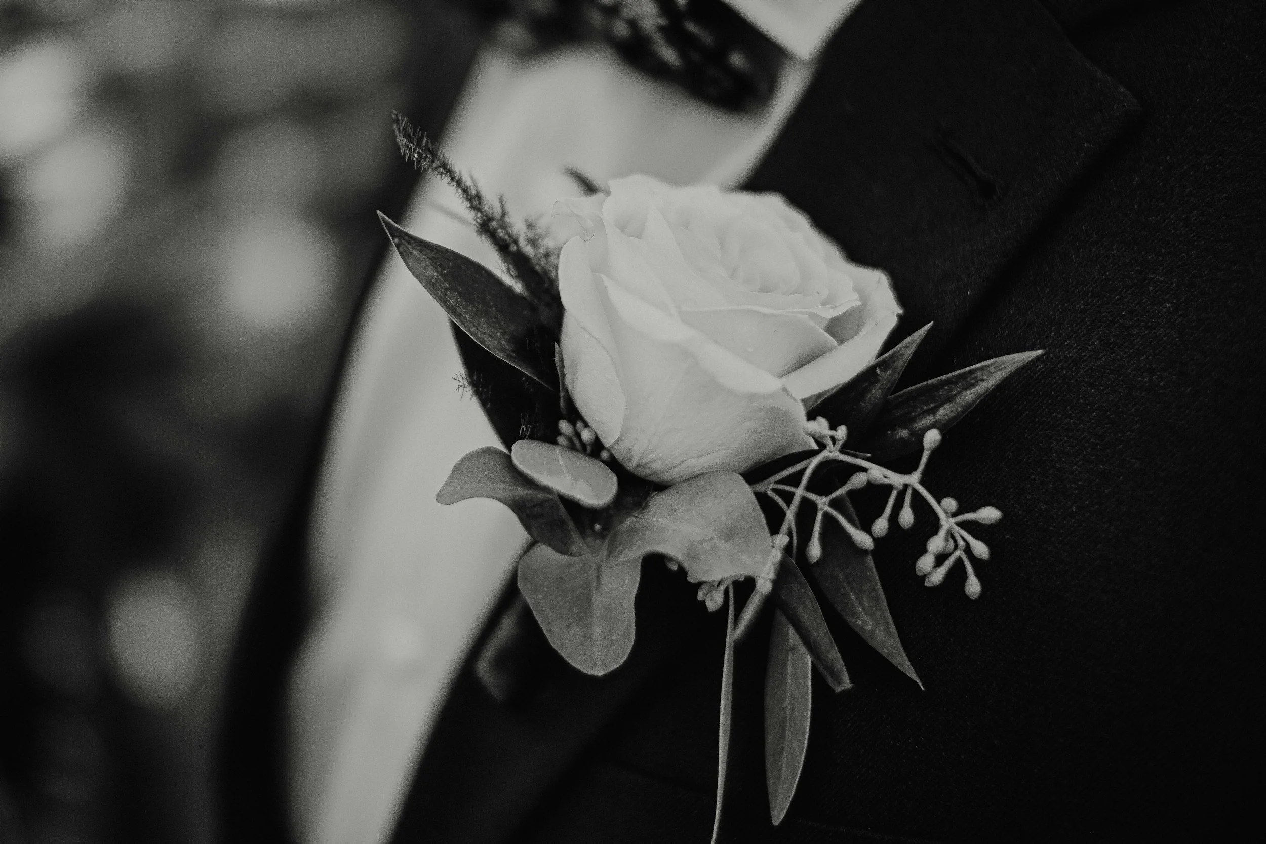 Black and white photo of a person in a suit with a boutonniere featuring a rose and greenery pinned to their lapel.