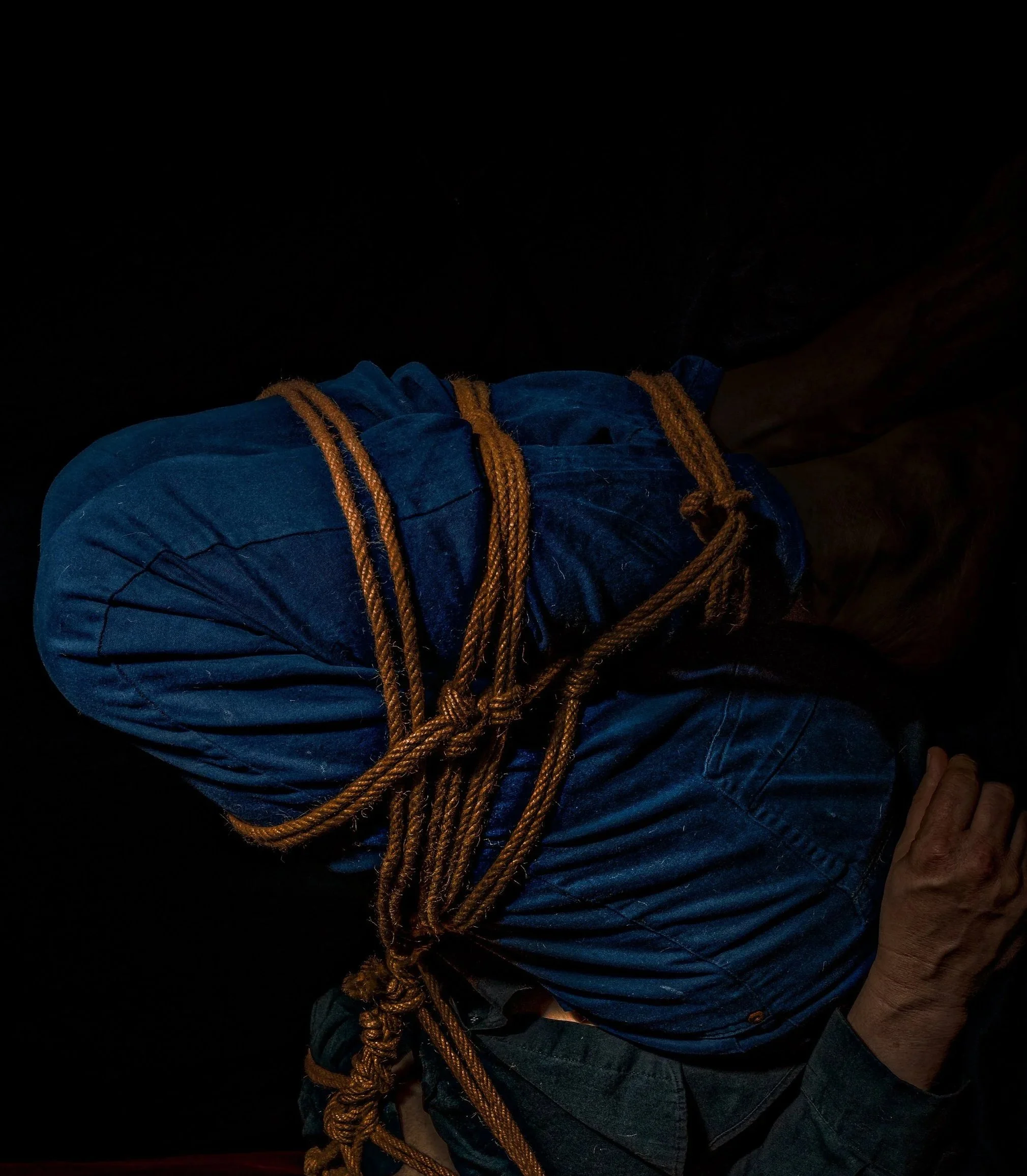 A person kneeling with their arms crossed, tied with orange ropes against a dark background.