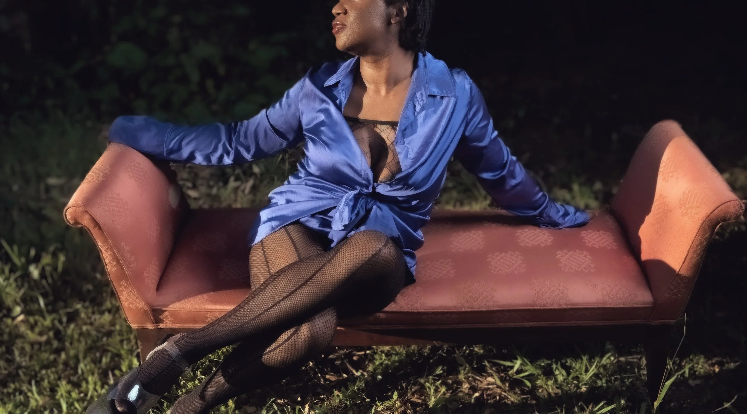 A woman wearing a blue satin shirt and fishnet stockings is sitting on an orange vintage sofa outdoors at night, with her head turned to the side and one arm resting on the sofa arm.  Intimate Portrait photography.