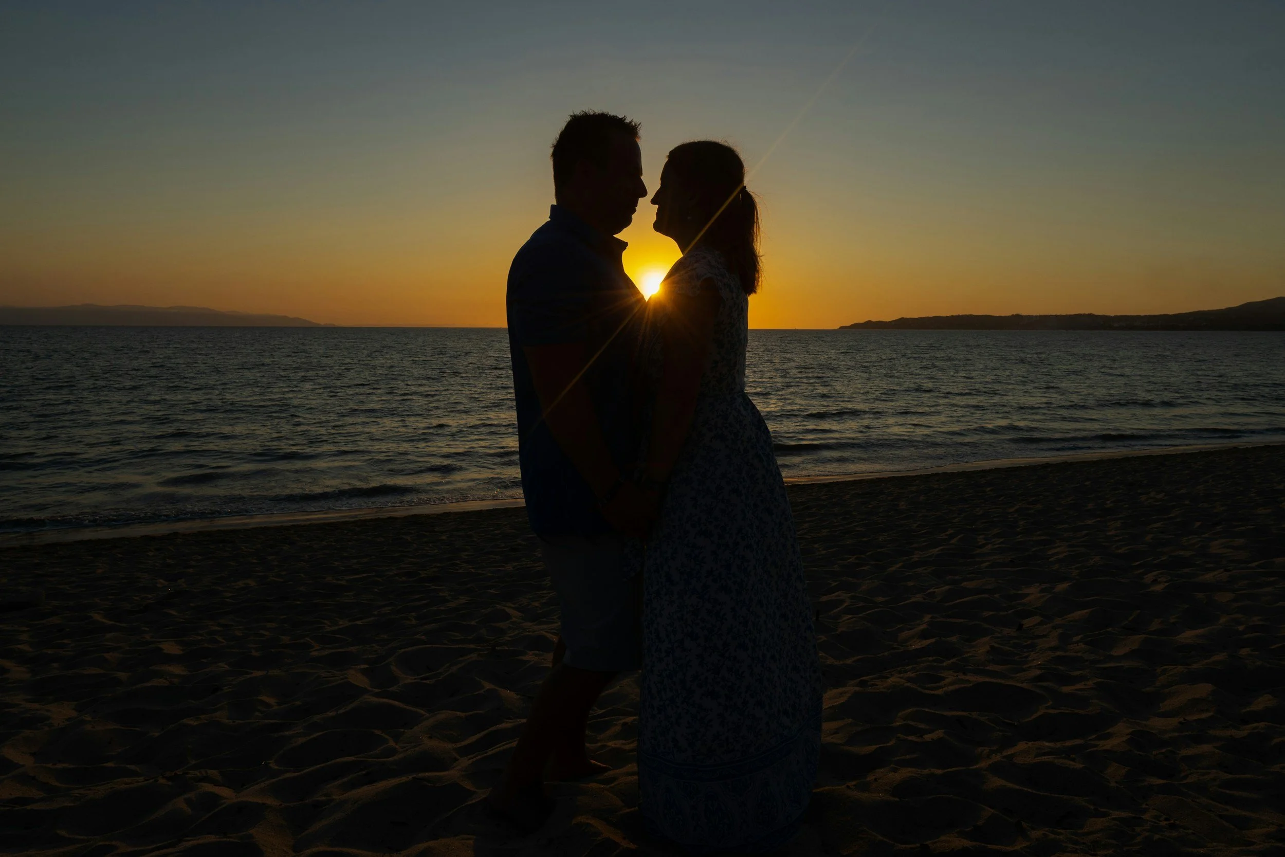 Silhouette of a couple standing on the beach at sunset, facing each other with the sun setting over the water in the background.