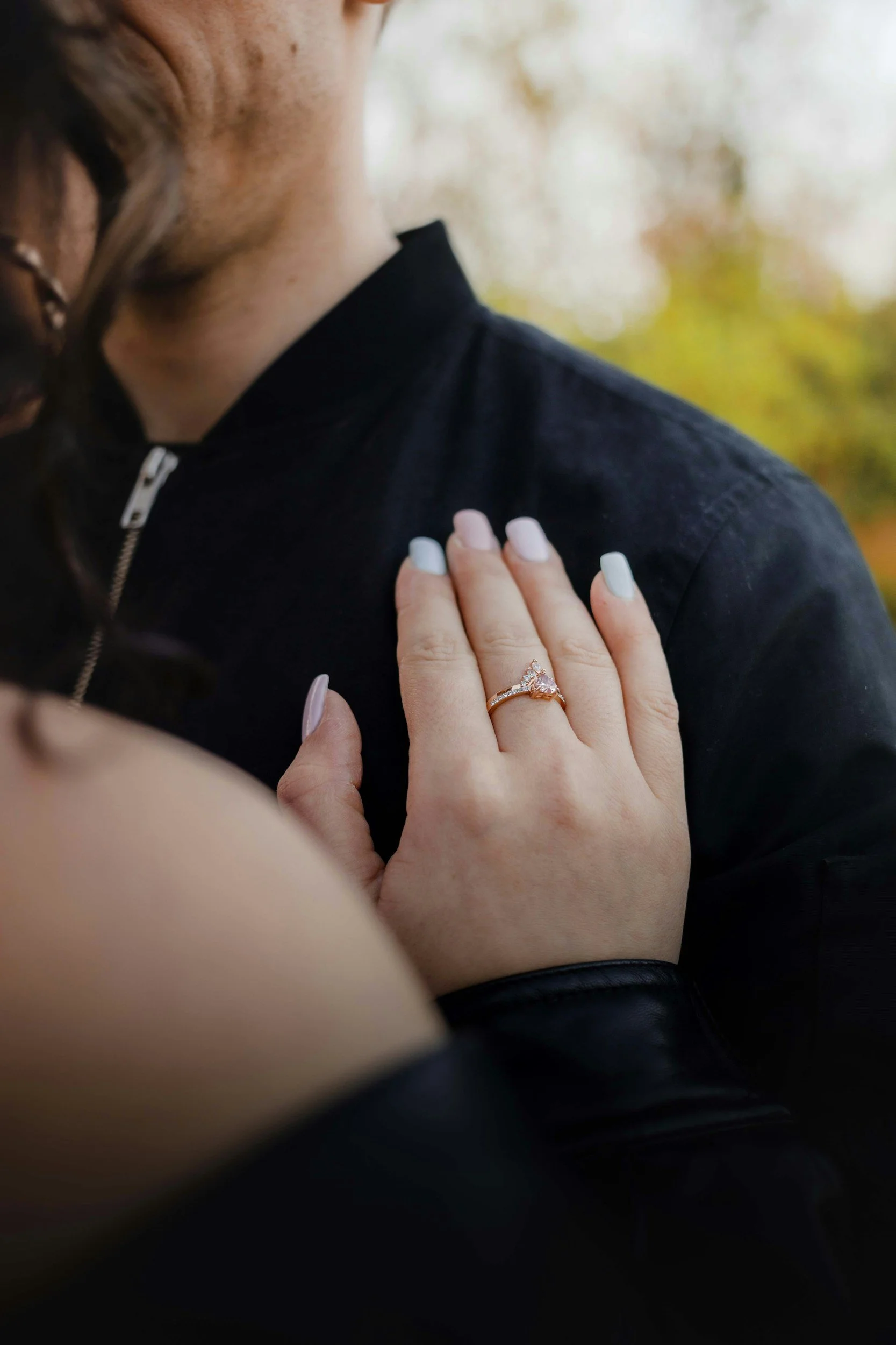 A woman’s hand with a ring resting on a man’s chest. The woman has light-colored nails and is wearing makeup. The man is wearing a black jacket with a zipper, and the background is blurred with autumn foliage.