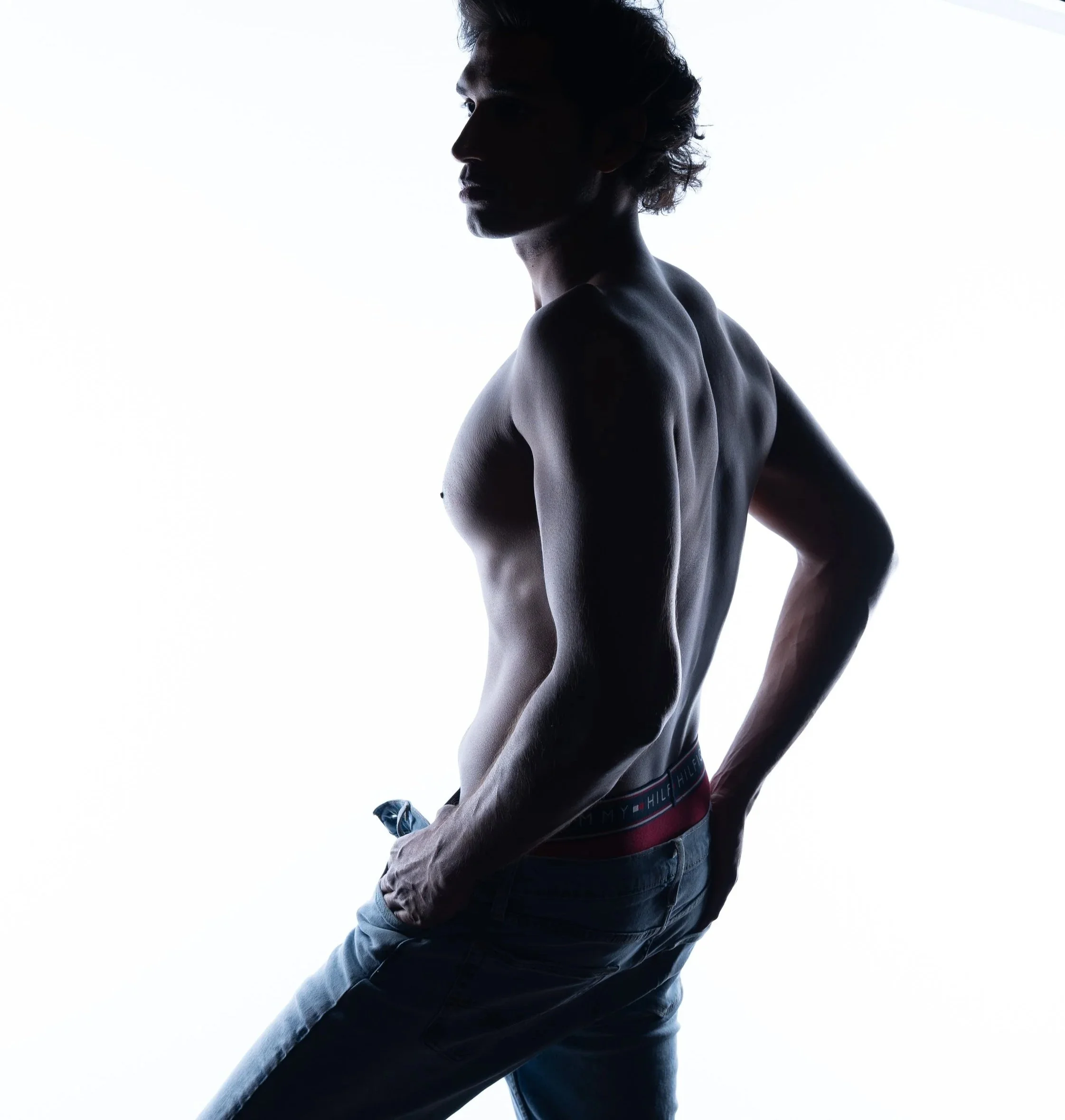 Silhouetted young man with tousled hair looking to the side, wearing jeans and a Tommy Hilfiger branded waistband, standing against a plain white background.  Boudoir for Men.  Male Boudoir. Gentlemen Boudoir.  Dudeoir and masculine boudoir.