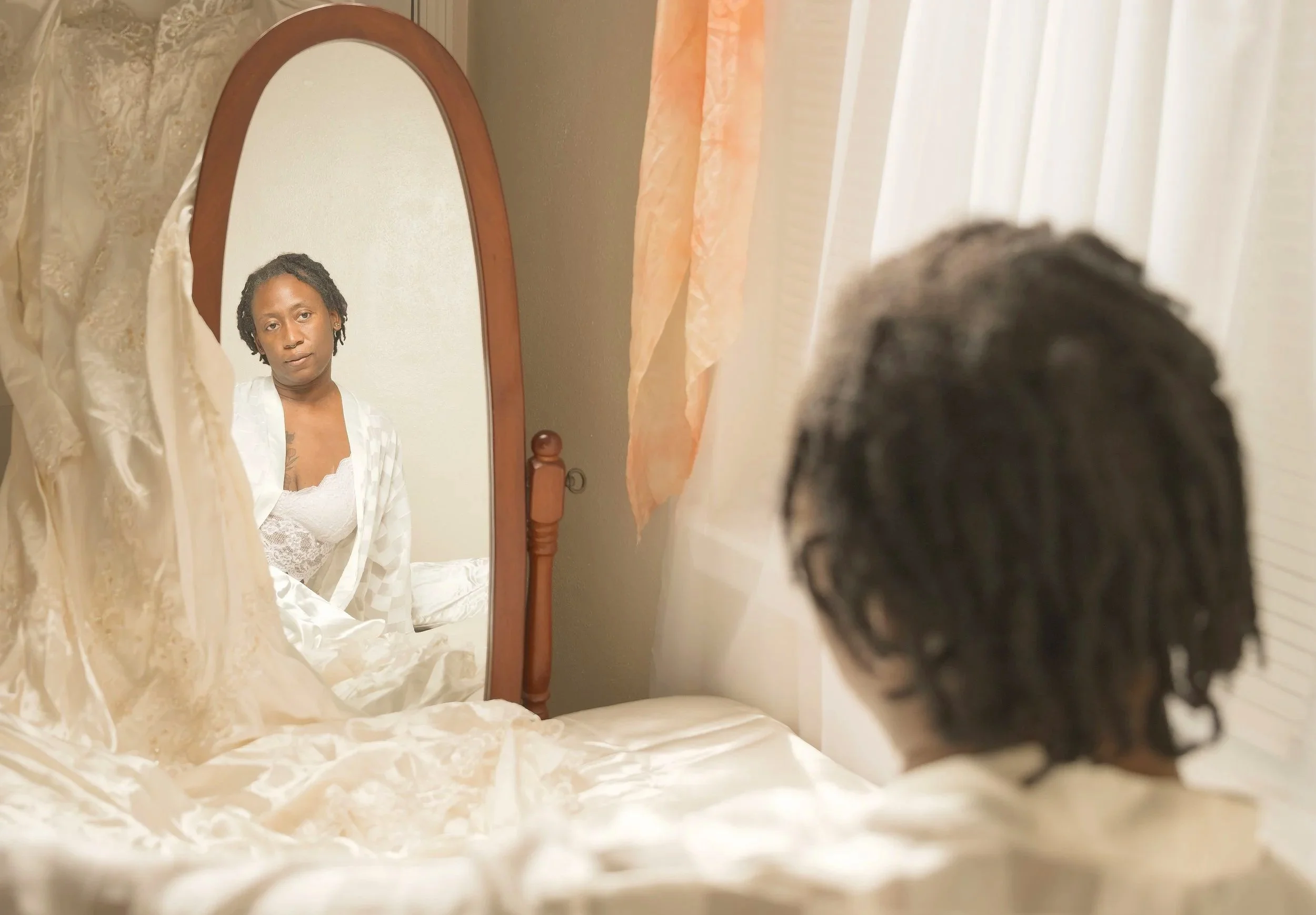 A bride with short curly hair looking into a mirror. She is wearing a white lace top and a light-colored satin robe, sitting on a bed with cream-colored sheets and a decorative pillow. Her wedding dress is hung next to the mirror.