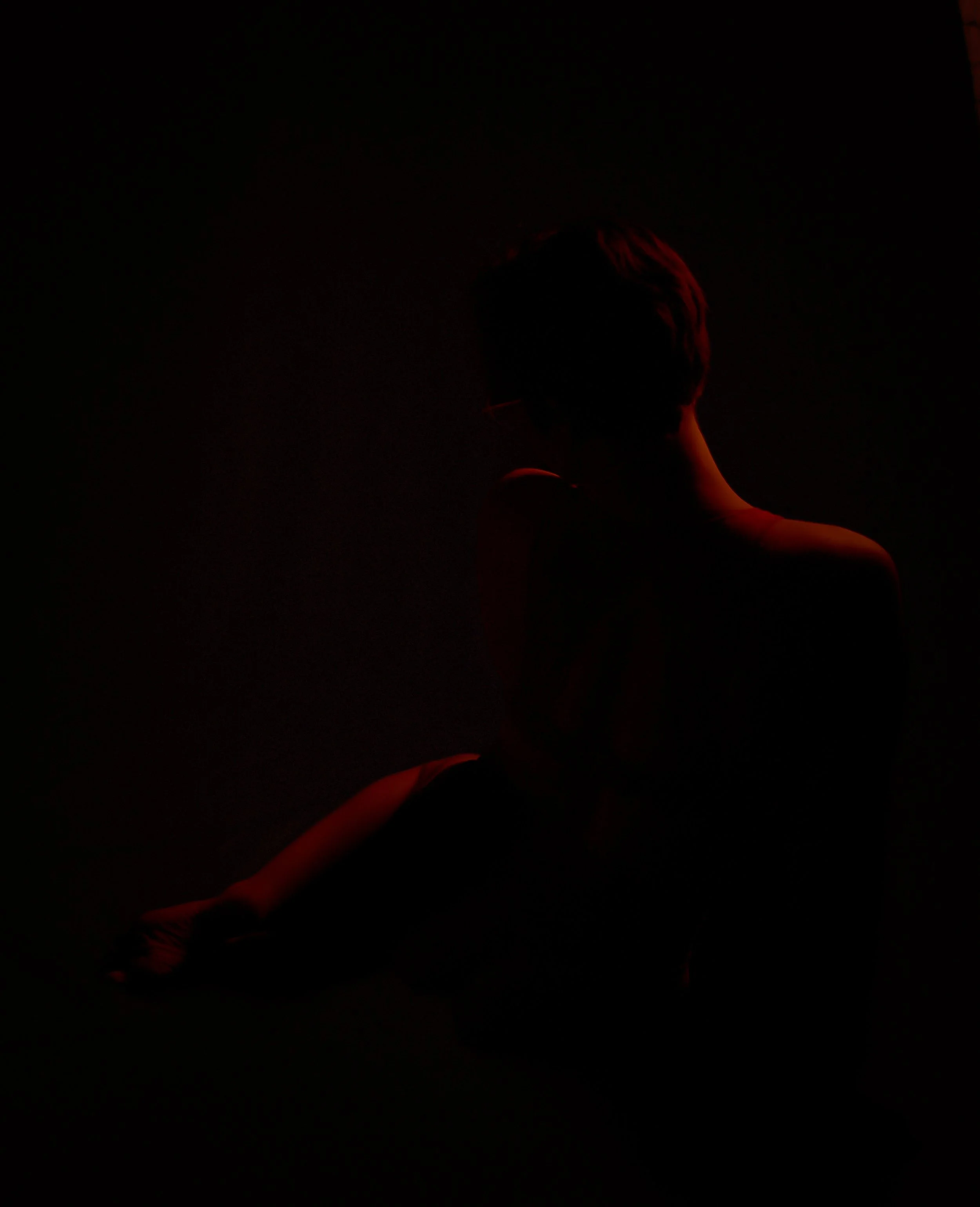 A person with short hair sitting in darkness with a faint red light illuminating their shoulder and arm.