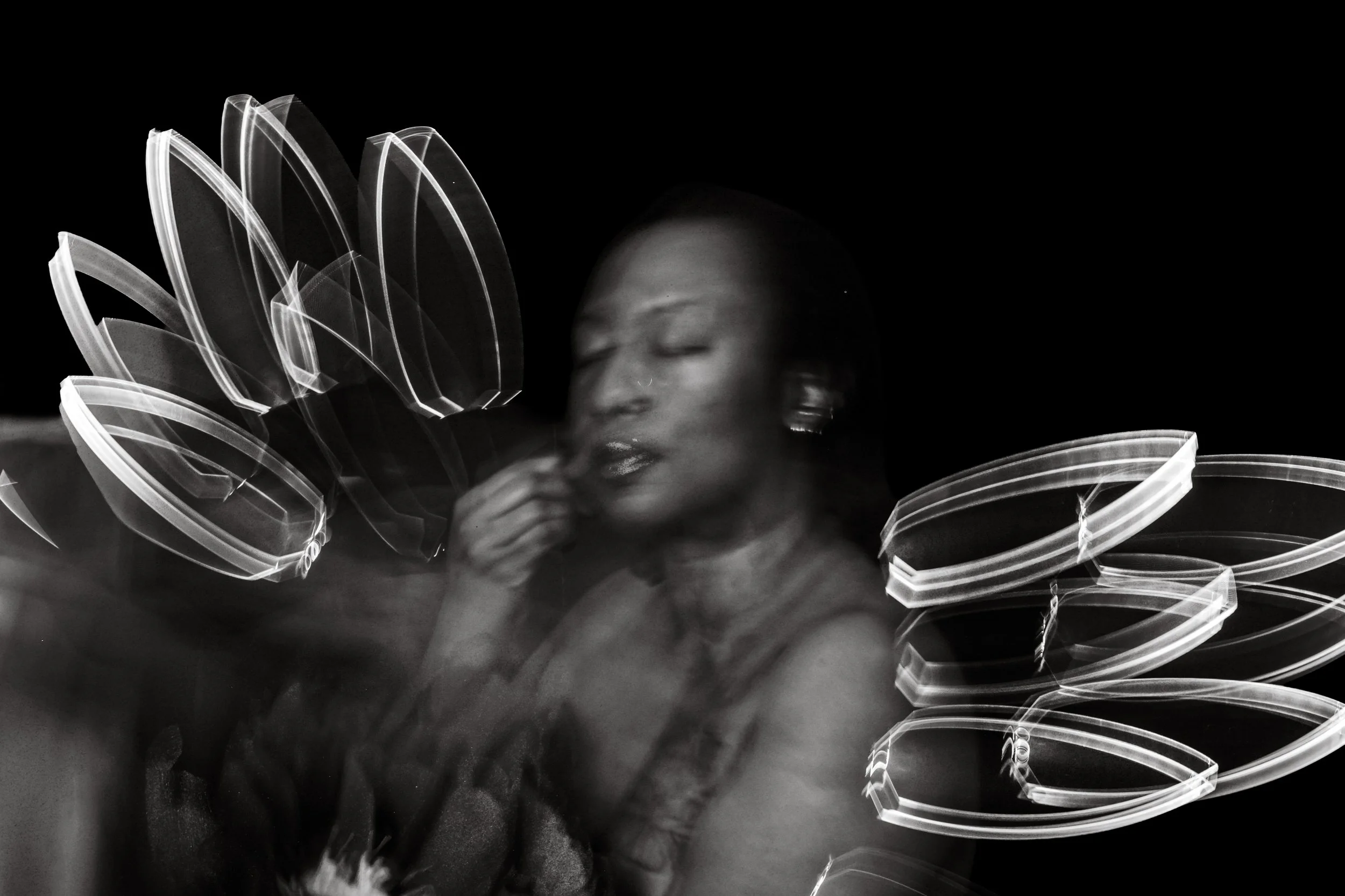 Painterly black-and-white light painting portrait of a woman with eyes closed and hand near her lips, framed by sharp geometric white light trails against pure black, Bellissimo Expressions studio.