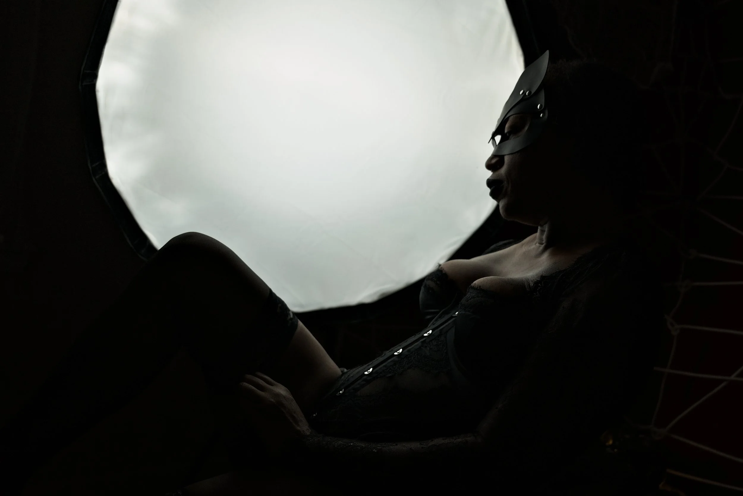 Silhouette of a woman wearing a bunny mask and black lingerie, sitting inside a dark tent or enclosure with a large white light or backdrop behind her.