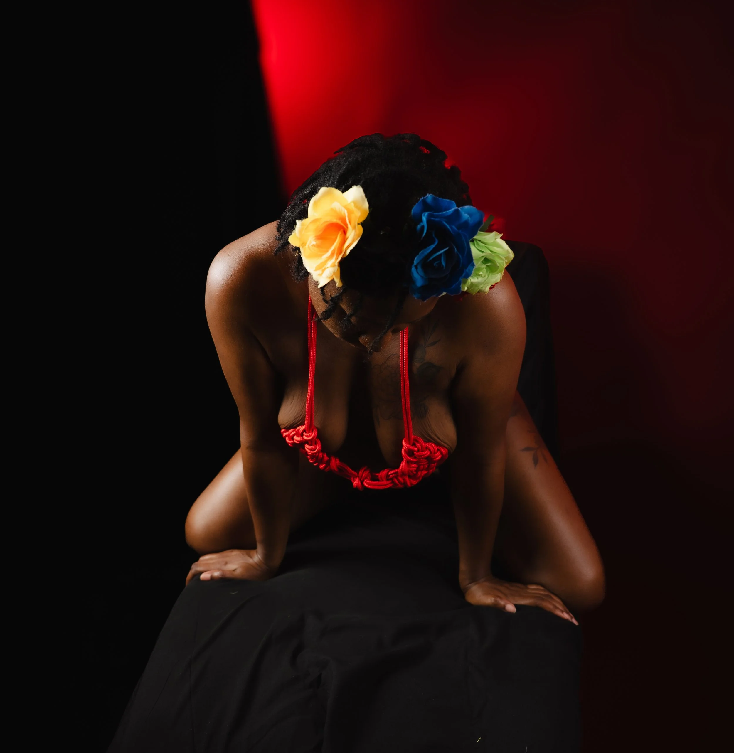Shibari portrait of a kneeling woman with a vibrant yellow and blue floral crown and red rope chest harness, head bowed, against a split black and red background.