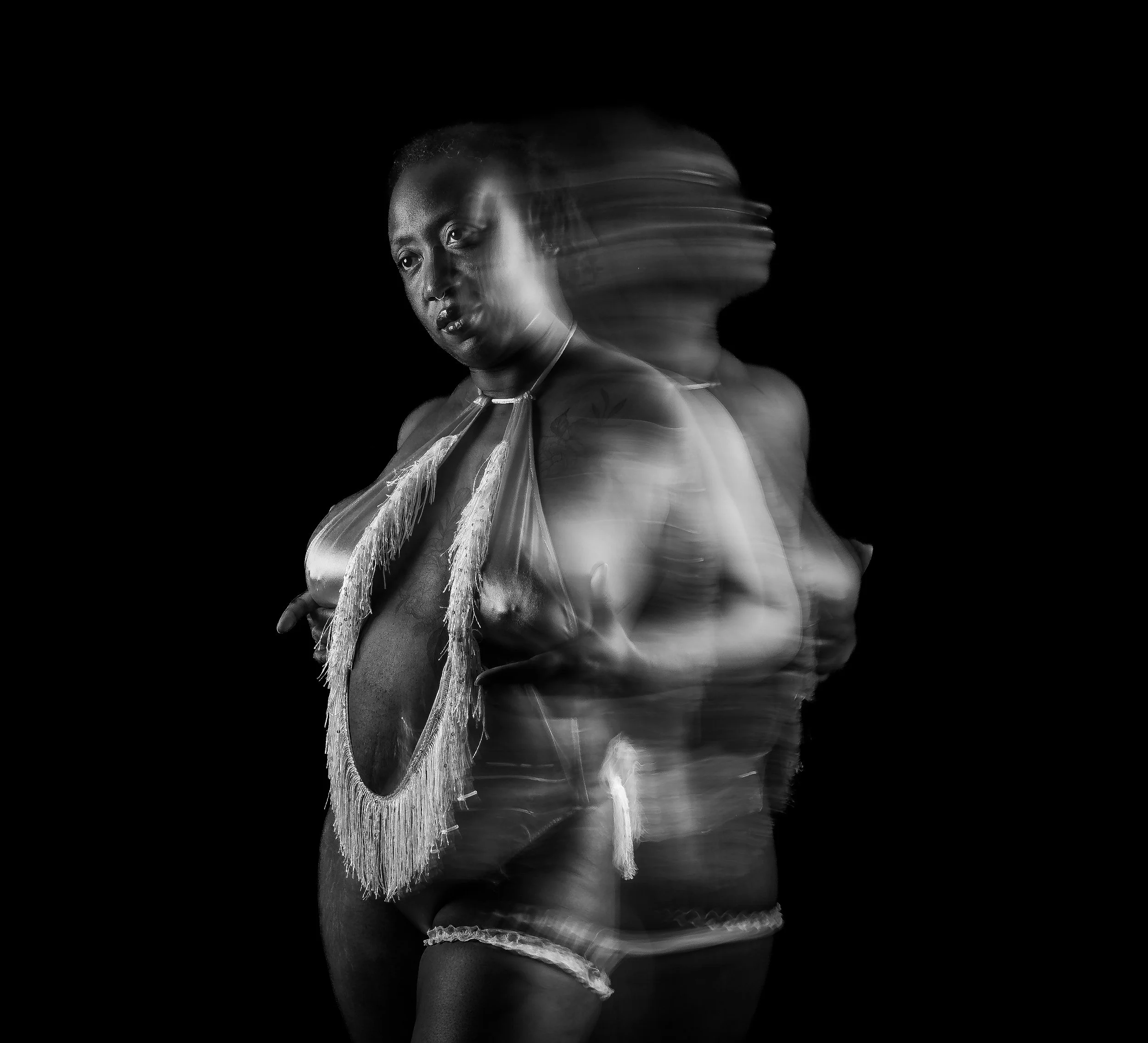 Black and white photo of a woman with motion blur creating a double exposure effect, wearing a beaded necklace and minimal clothing.