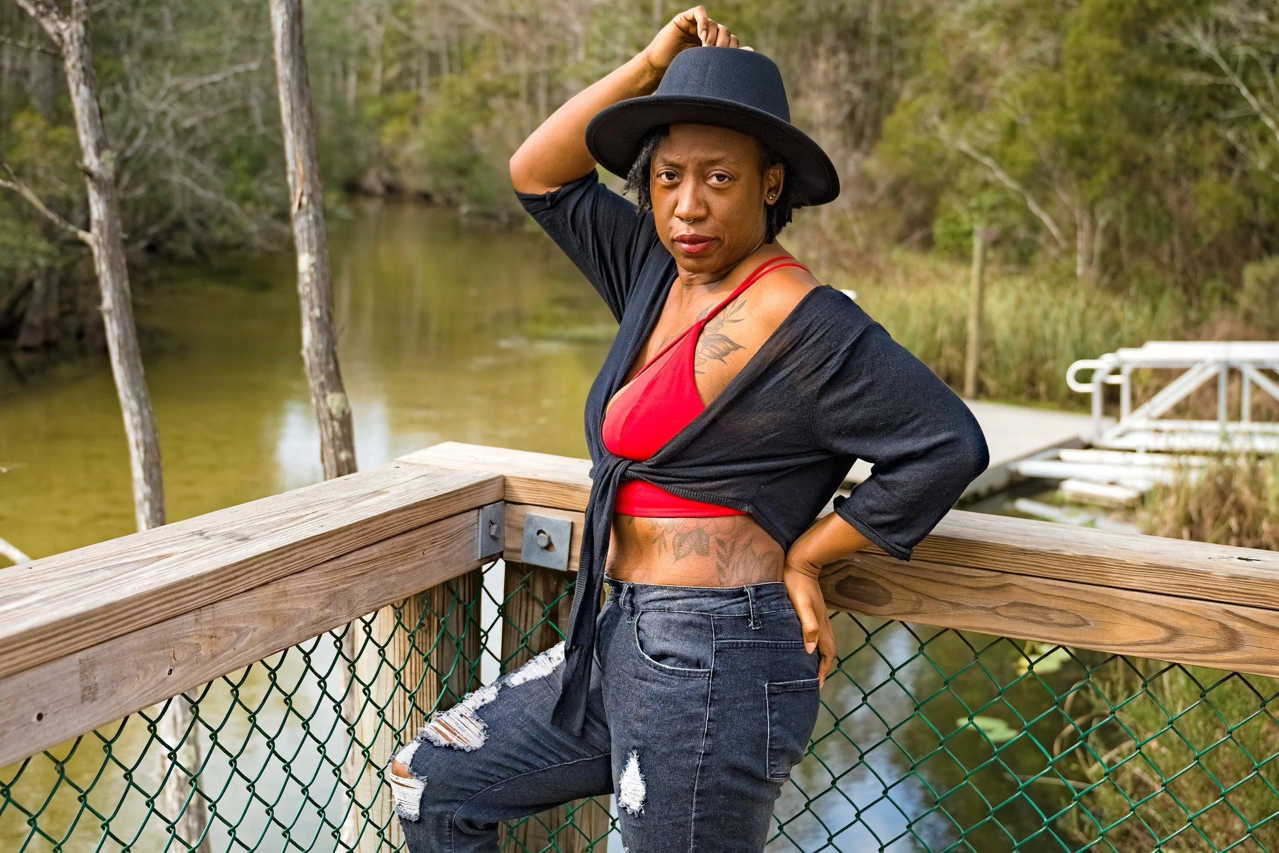 A woman standing on a wooden bridge near a body of water, wearing a black hat, red top, black cardigan, and ripped black jeans.