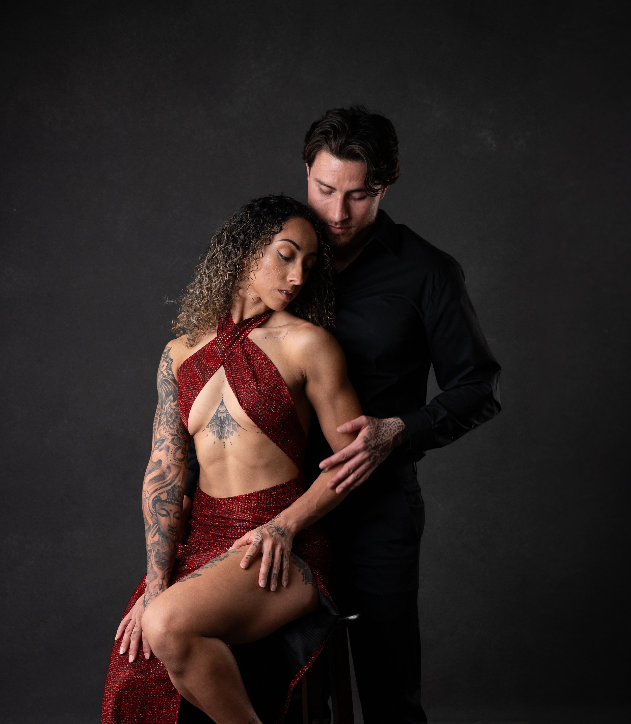 A man and a woman pose intimately against a dark background. The woman is dressed in a red, glittering dress, and has curly hair and visible tattoos on her arm. The man, dressed in a black shirt, stands closely with his head bowed, touching the woman
