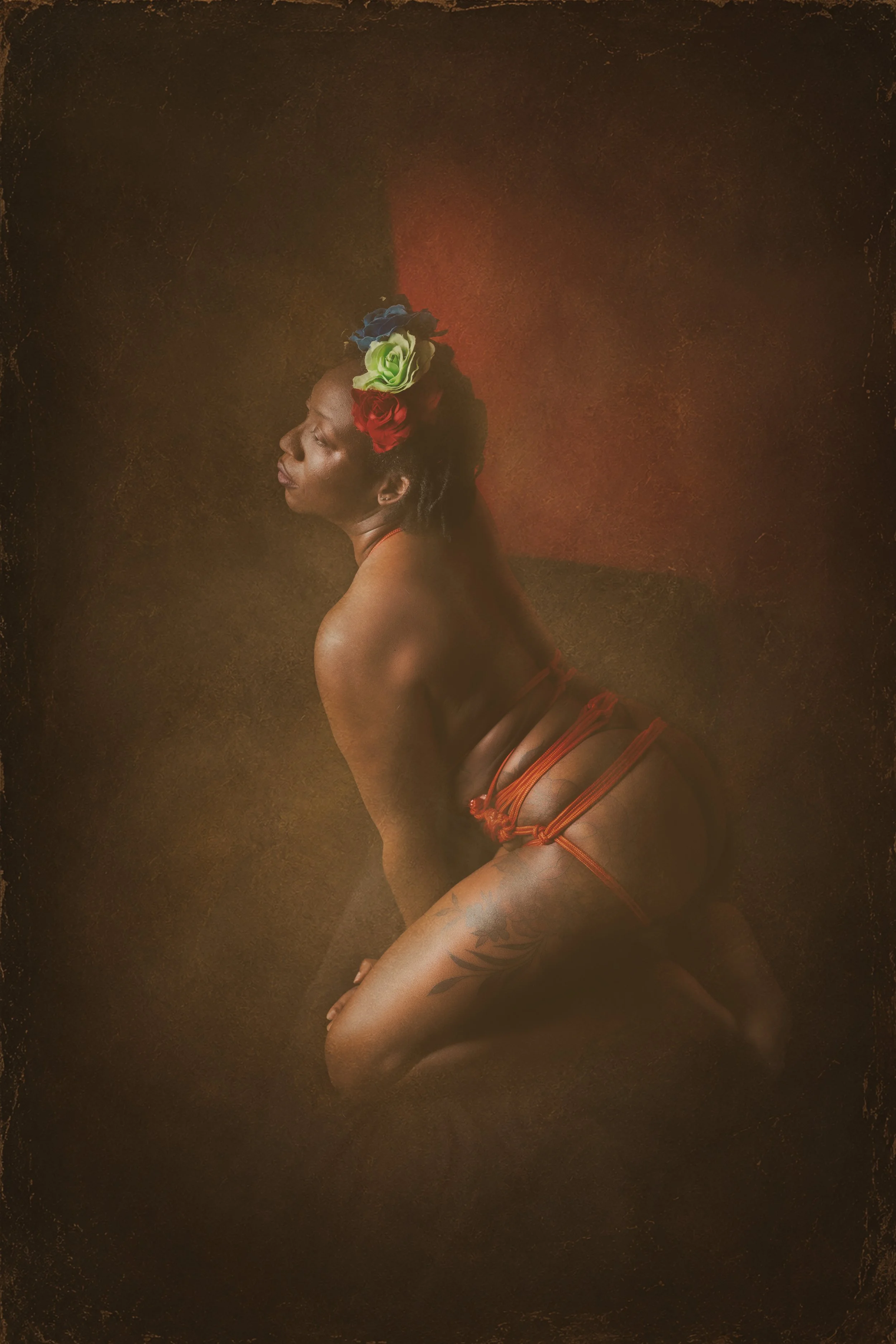 A woman tied in red shibari rope around her waist with a dark complexion, eyes closed, and head tilted back, wearing a large colorful flower crown and intricate red body art, against a dark, textured background.
