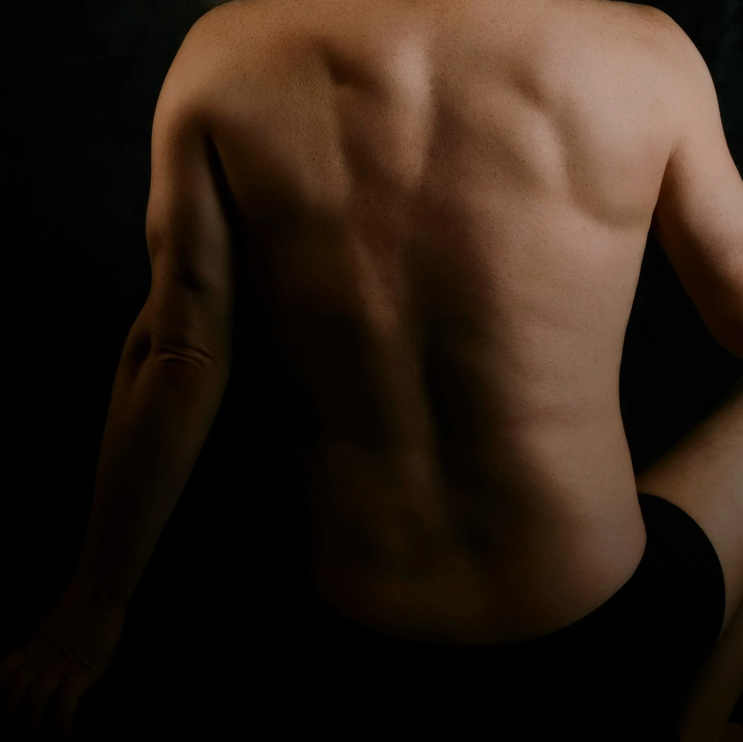 A black-and-white photo of a person's muscular back and arms, viewed from behind, wearing dark underwear. The dark background emphasizes the contours and strength of the pose. Beauty captured by Camera, Boudoir, weddings, Photography Gulf Coast, Emer