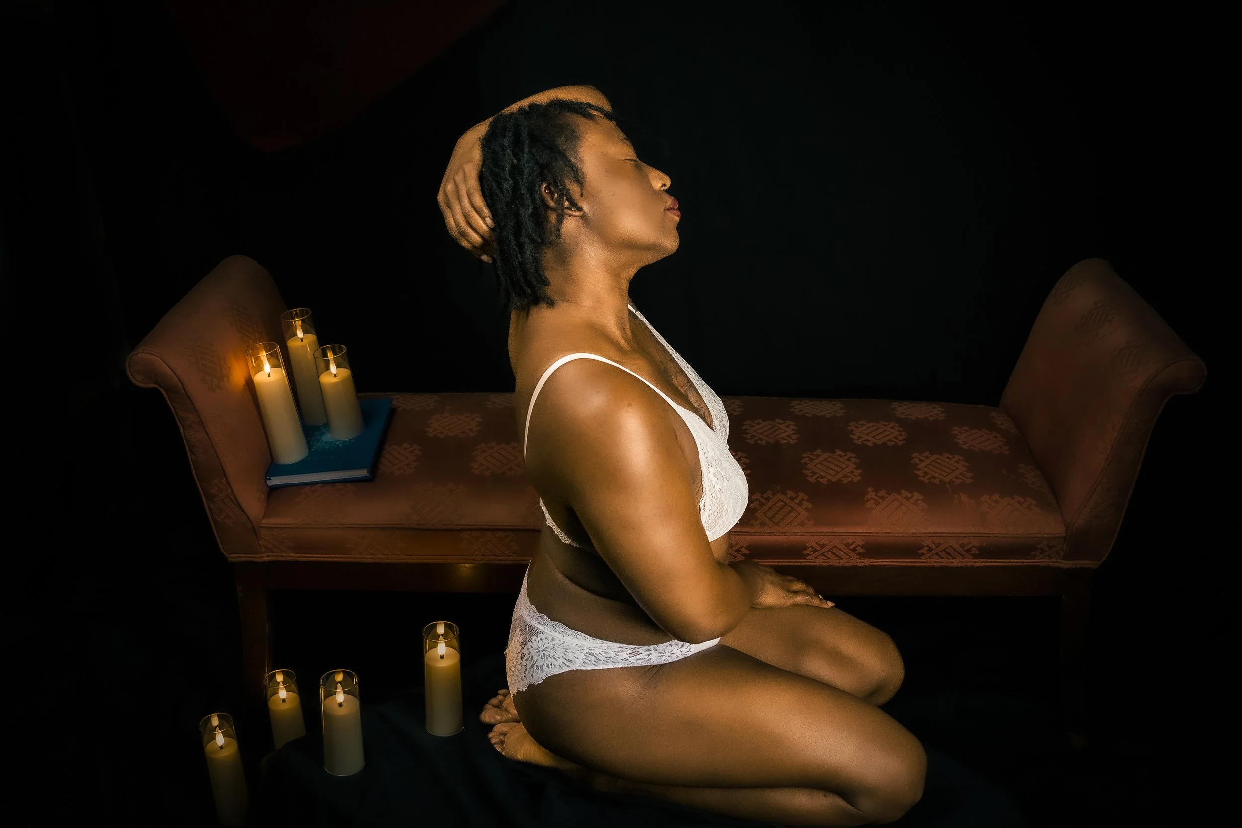 A woman in white lace lingerie kneeling on a black cloth in front of a vintage-style pink upholstered bench with candles on the seat and on the floor around her.