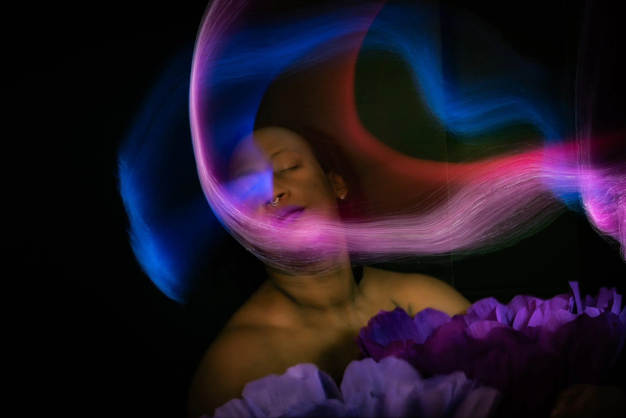 Artistic light painting boudoir portrait by Bellissimo Expressions – woman illuminated by glowing pink, blue and purple light trails with large peony flowers, Fort Walton Beach studio.