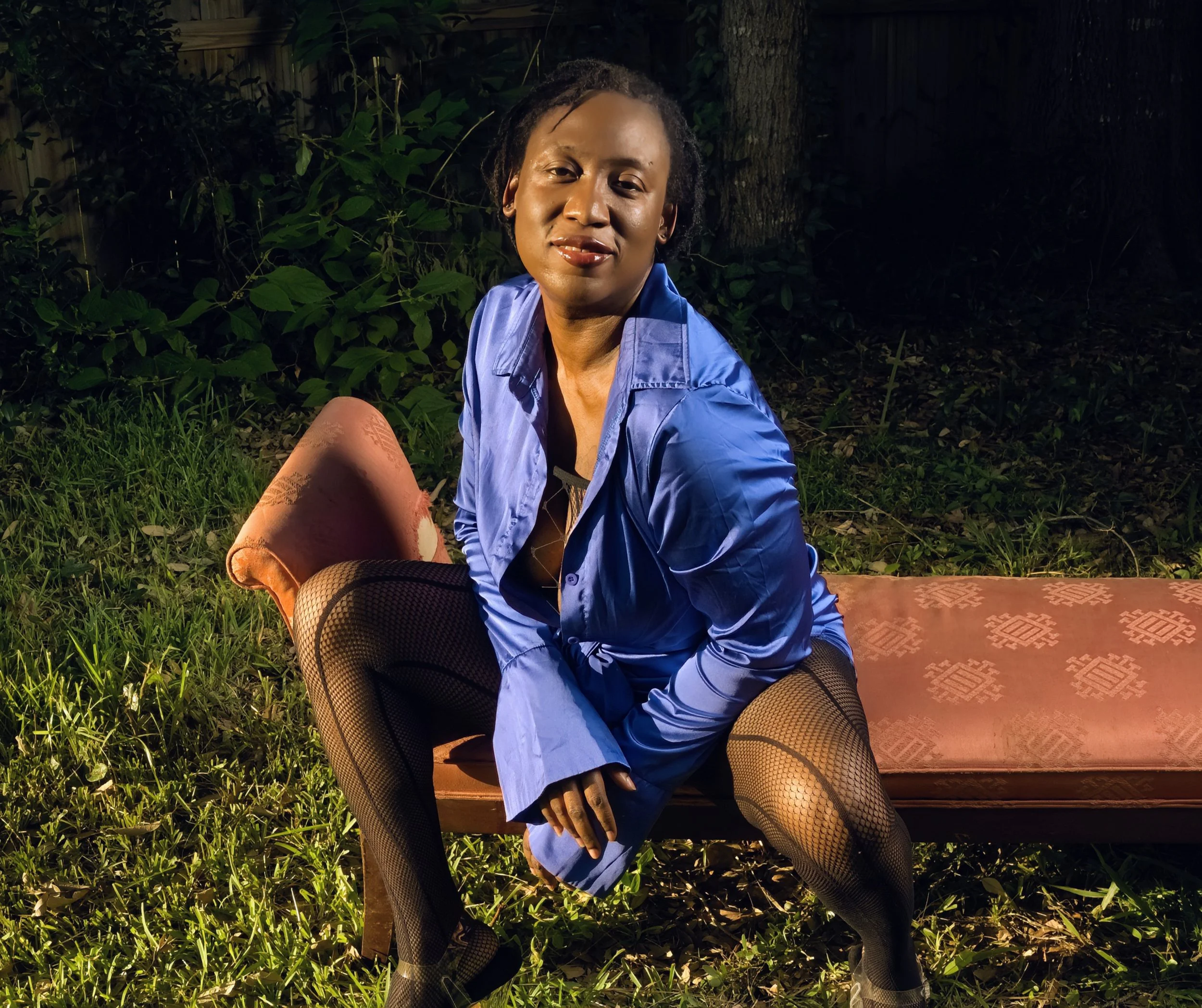 A woman sitting on an orange patterned bench outdoors at night, wearing a blue top and fishnet stockings leaning forward with smirky grin. Her blue top is unbuttoned, revealing a peak inside. The background features grass and trees, illuminated by s
