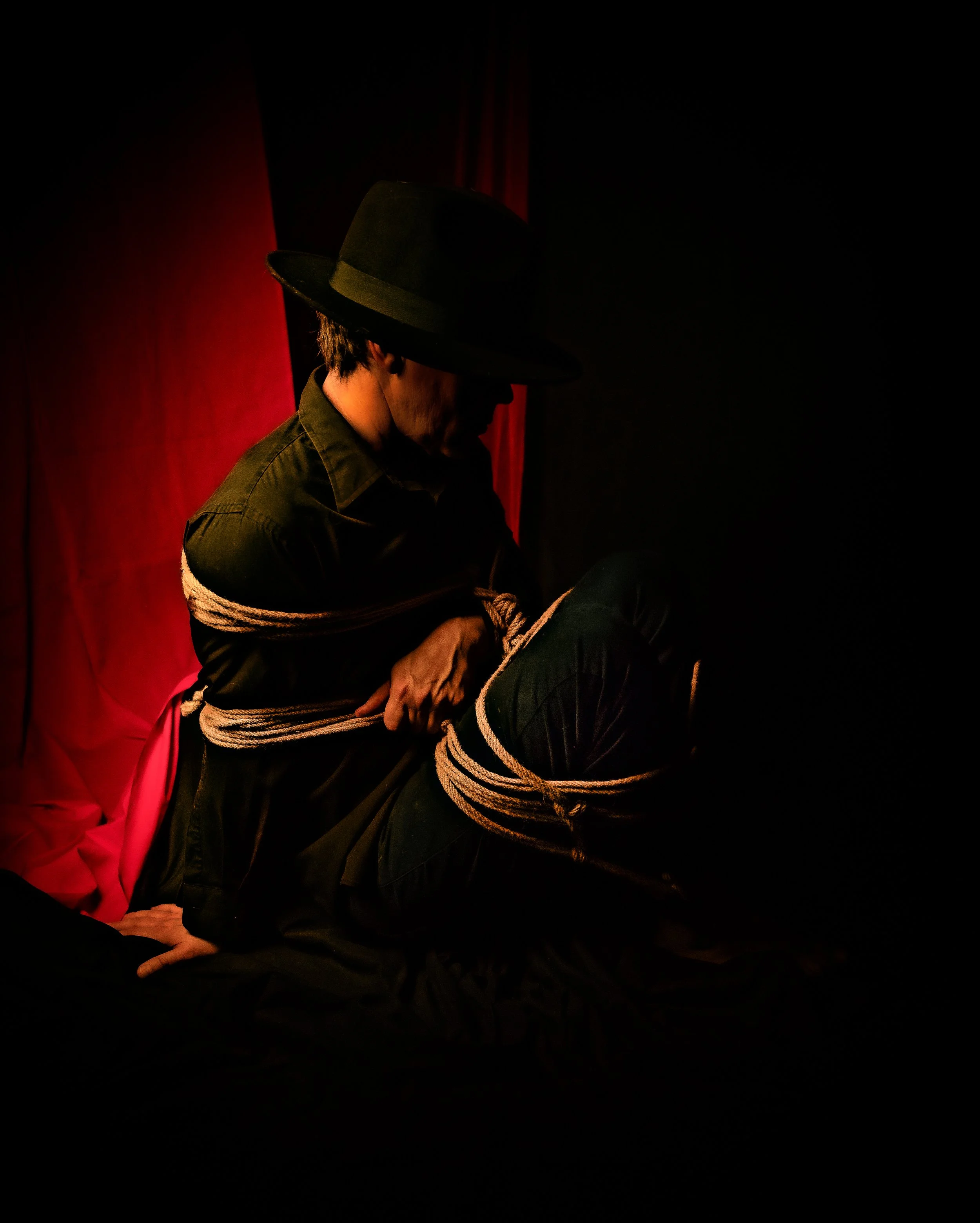 A person wearing a black hat and dark clothing is restrained with ropes in a dimly lit setting, with a red curtain in the background.