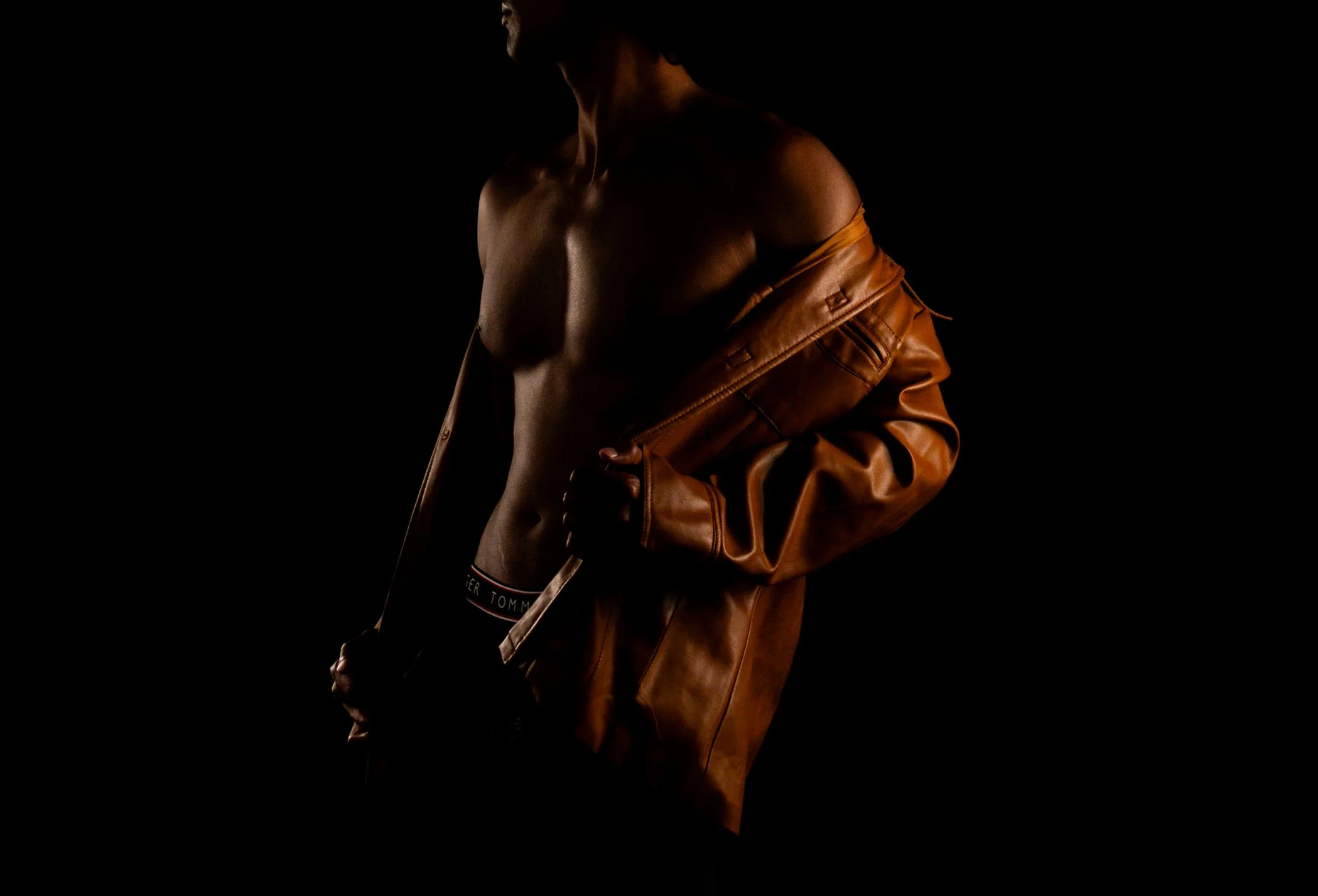 Moody studio portrait of a shirtless man holding an open tan leather jacket, lit with warm directional light against a black background.  Boudoir for Men.  Male Boudoir. Gentlemen Boudoir.  Dudeoir and masculine boudoir.