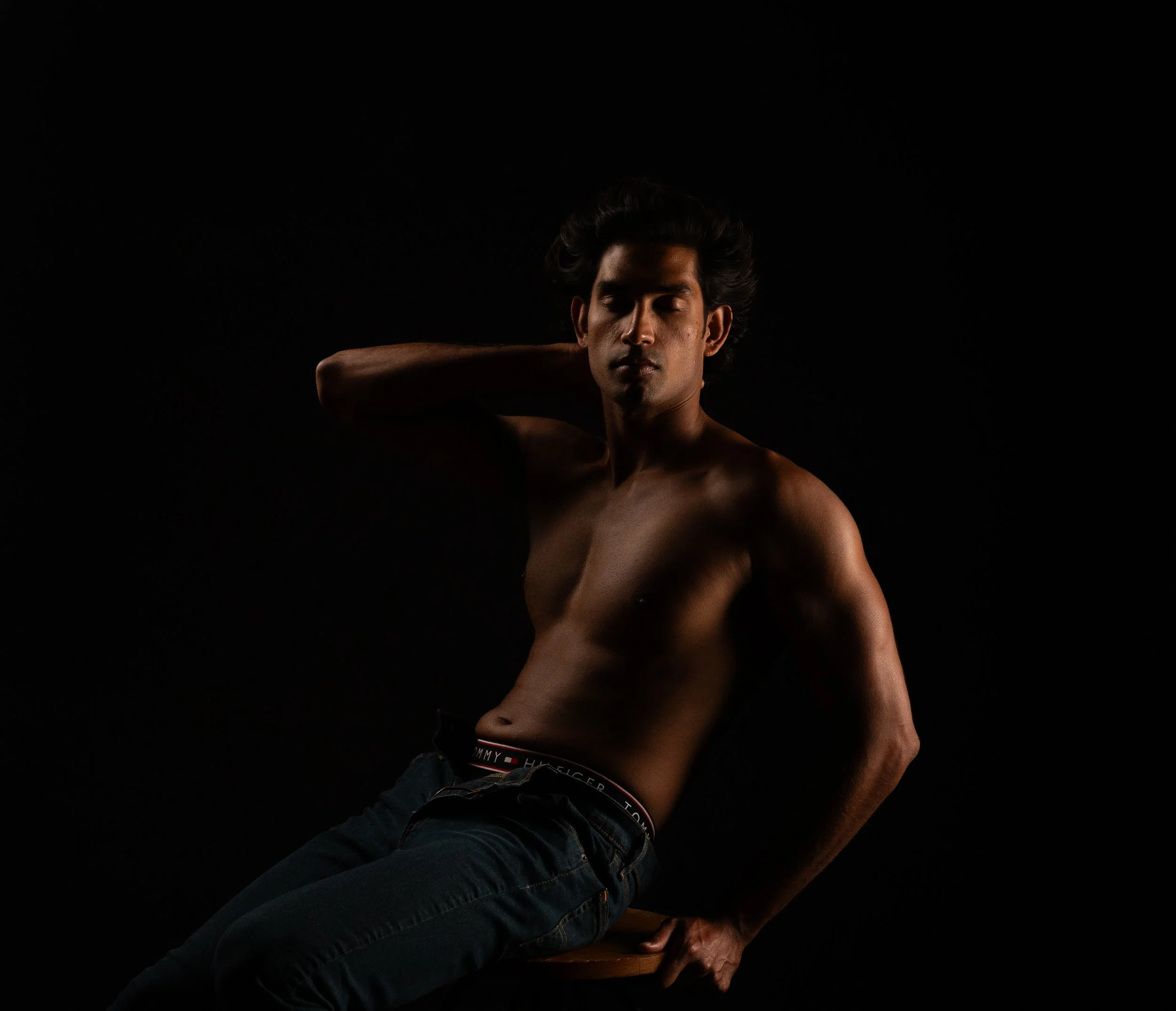 Dark, moody studio portrait of a shirtless man in jeans, hand raised near his face, lit with minimal low-key lighting against a black background.  Boudoir for Men.  Male Boudoir. Gentlemen Boudoir.  Dudeoir and masculine boudoir.