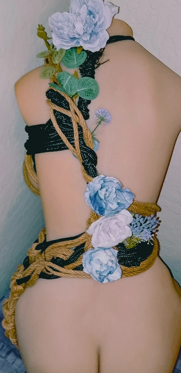 A person with their back turned, wrapped in decorative black and tan ropes, with artificial flowers attached to the ropes, creating a floral and rope art arrangement.