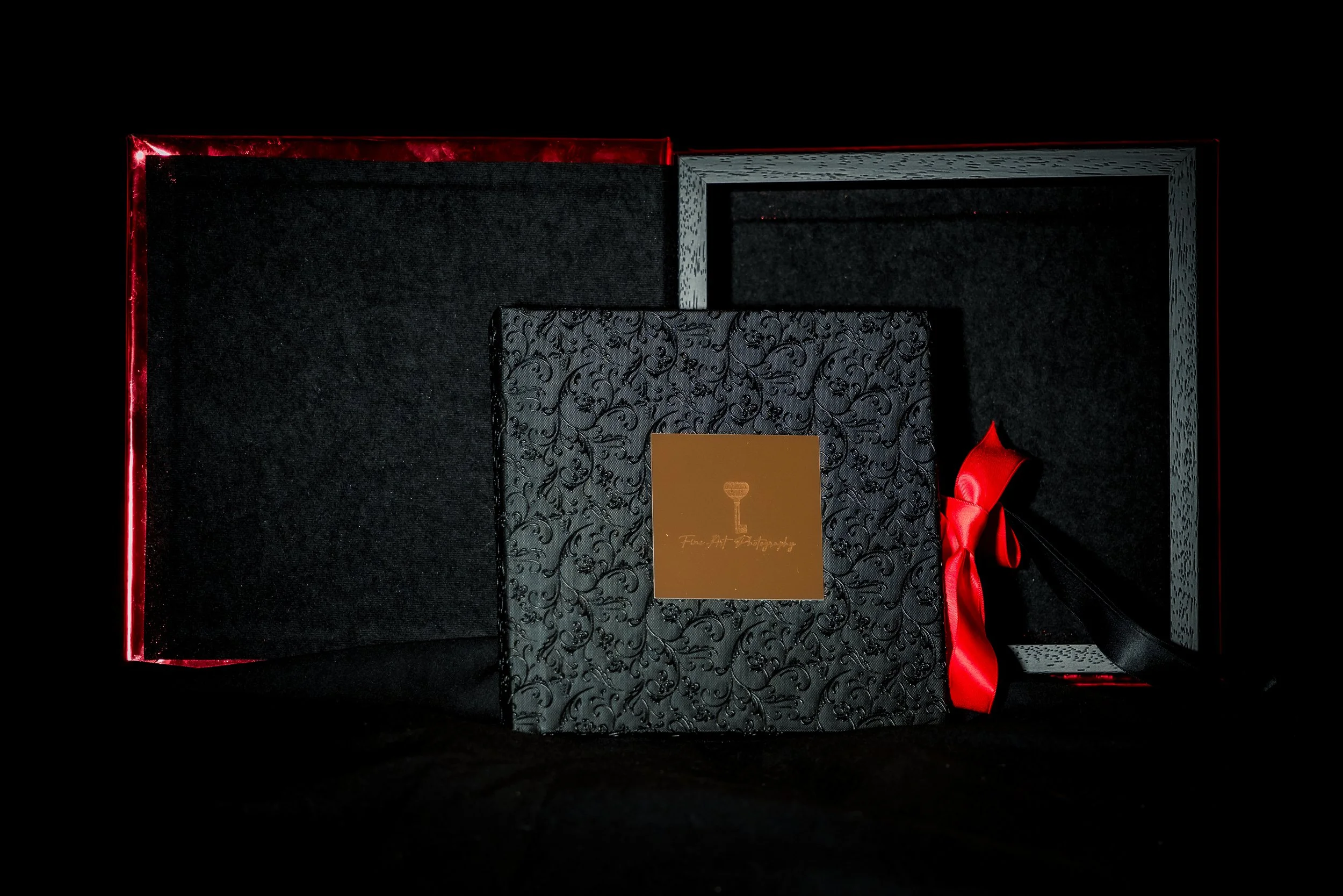 A black photo box with a red ribbon and decorative patterned black and gold card inside, surrounded by black felt fabric.