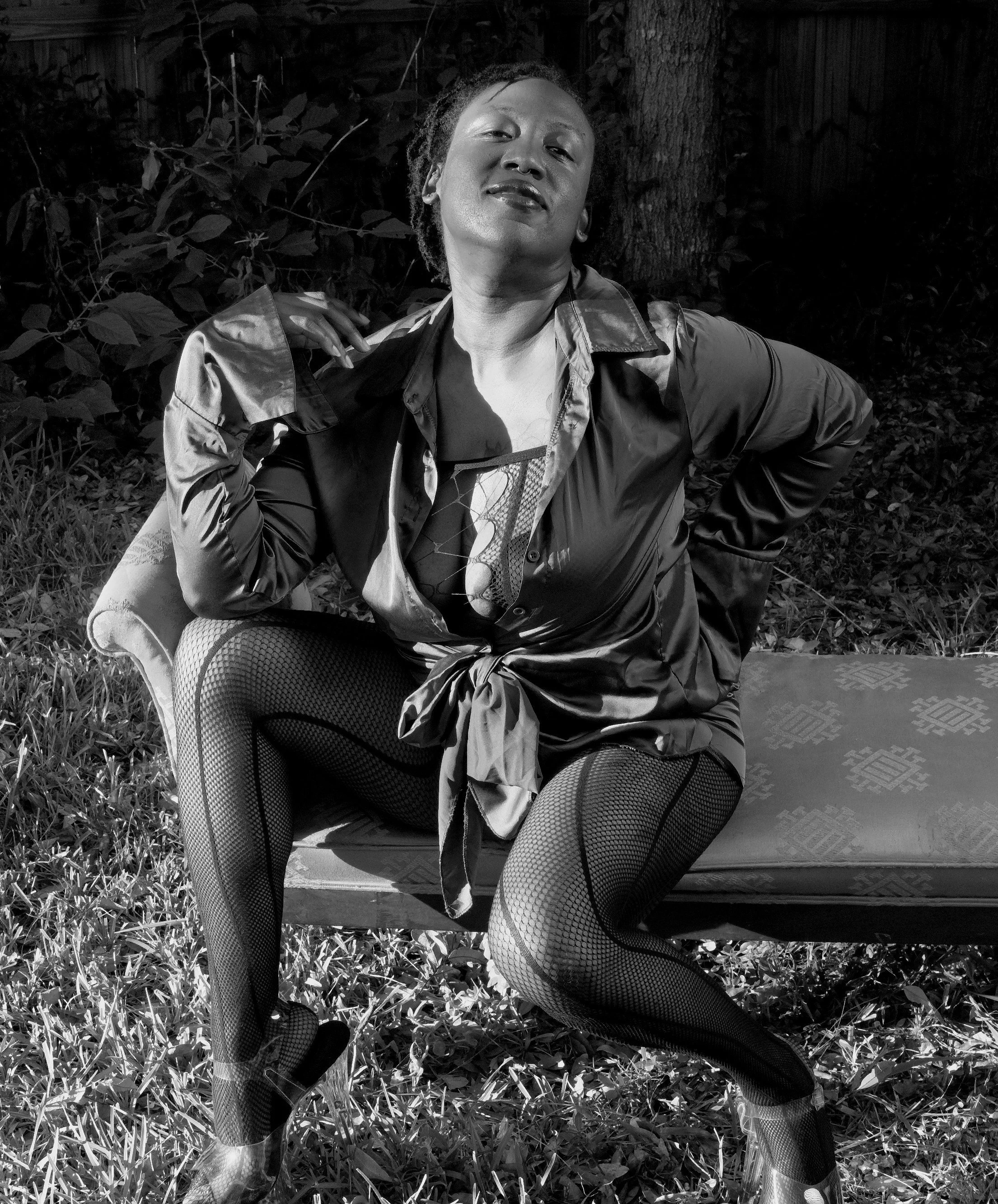 Lifestyle Boudoir. A black-and-white photo of a person sitting on a patterned bench outdoors, wearing a tied jacket, fishnet stockings, and heels. The background features grass and trees, adding a natural setting. Boudoir, weddings, Photography Gulf