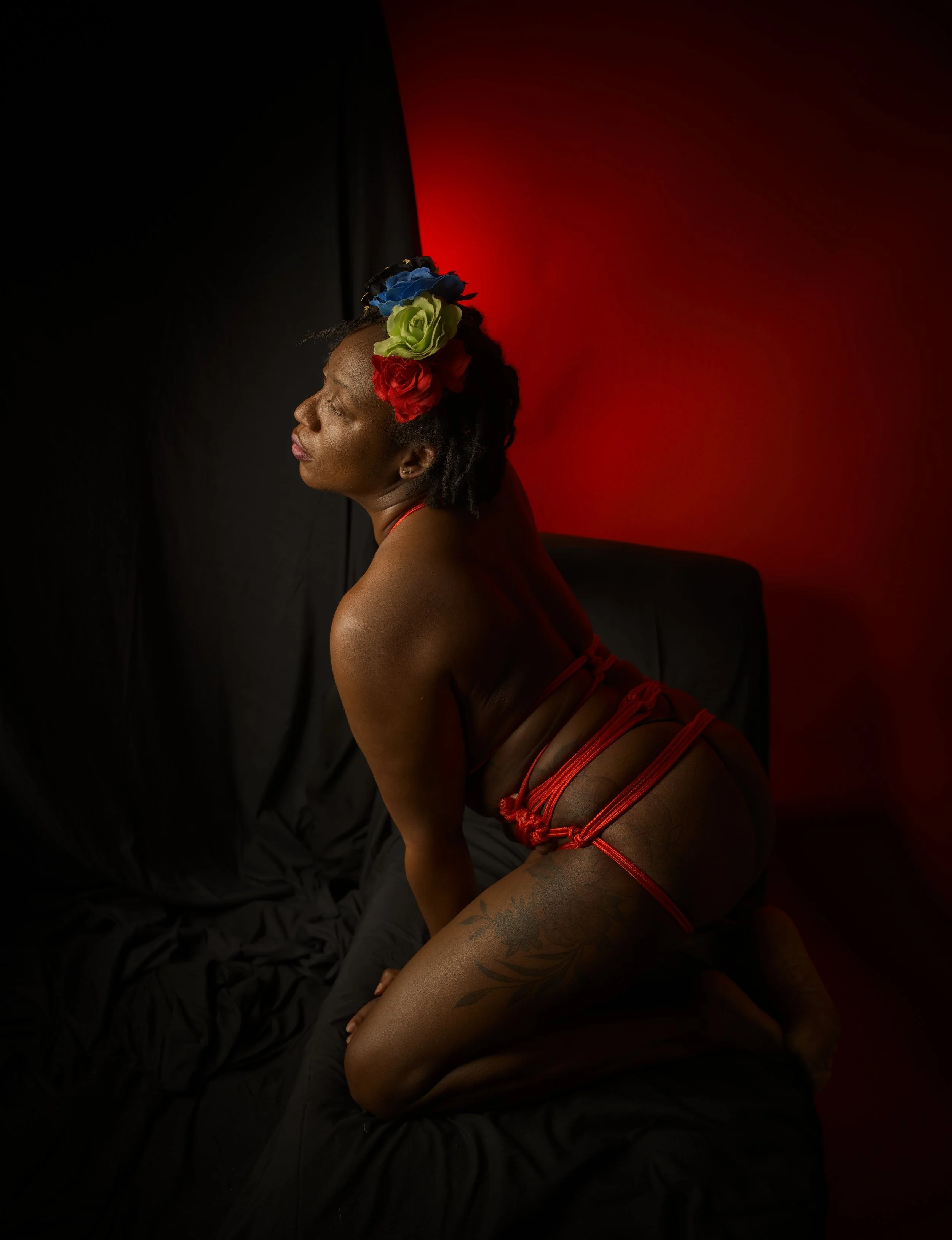 A woman sensually sitting on a black chair wearing red ropes around her waist and flowers in her hair.