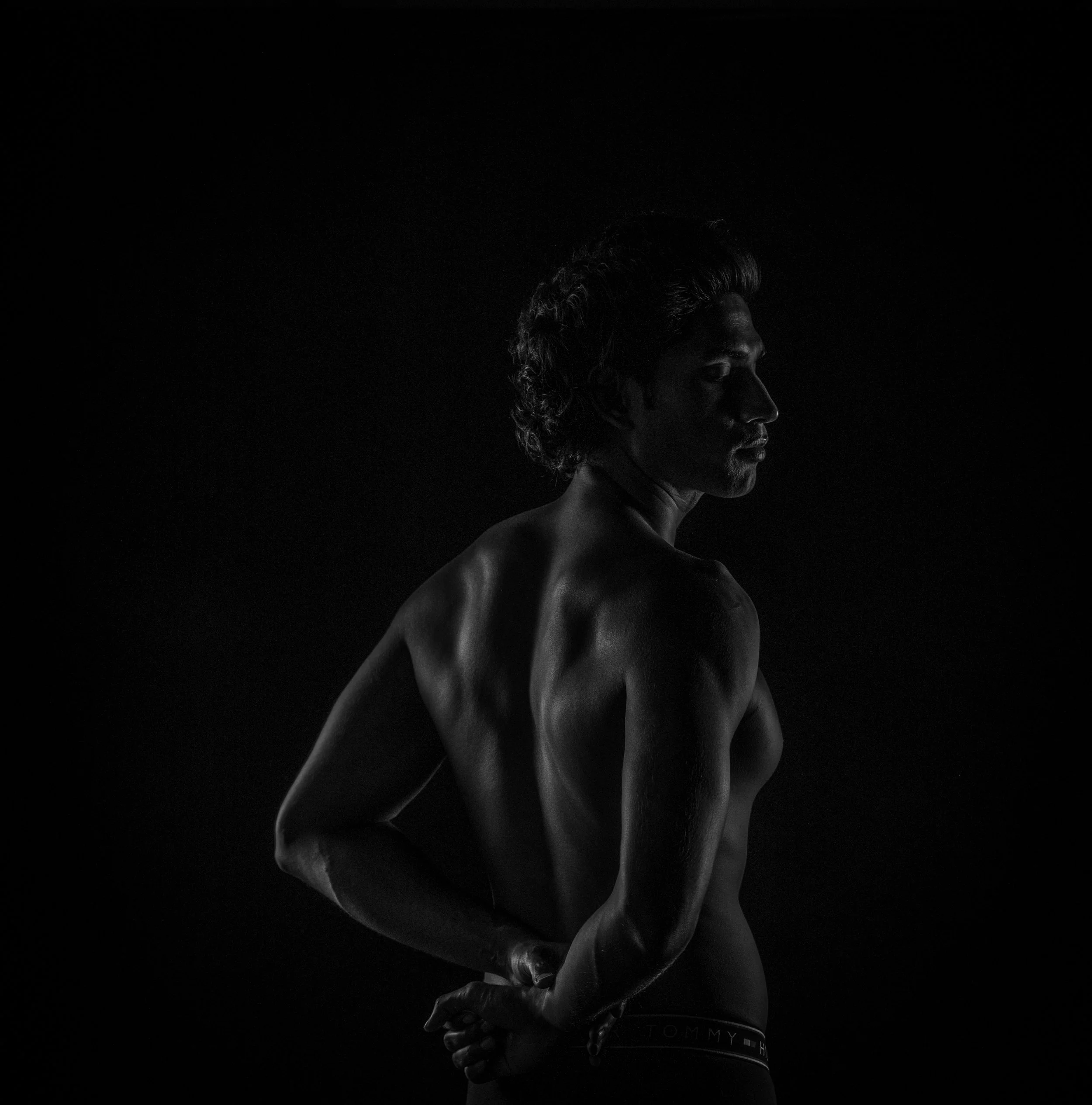 Low-key lighted photo of a black and white side profile of a man with curly hair, with her hands clasped in front of her, set against a dark background.
