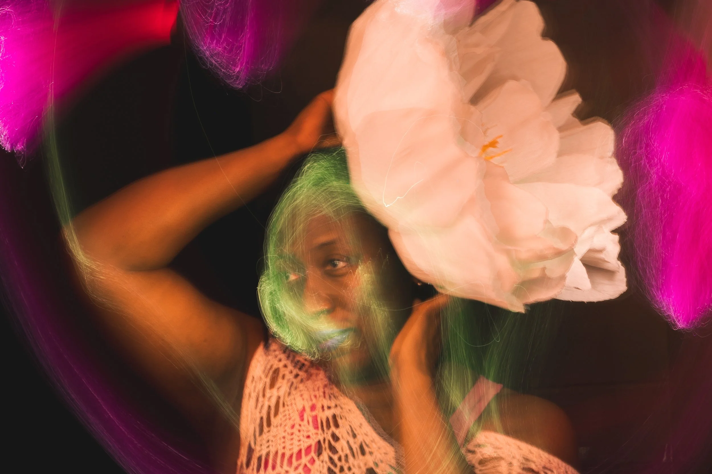 Painterly light painting portrait of a woman with arms raised framing a large pale pink flower against her head, green-tinted motion blur across her face and lace top, soft magenta light accents on a dark background, Bellissimo Expressions.