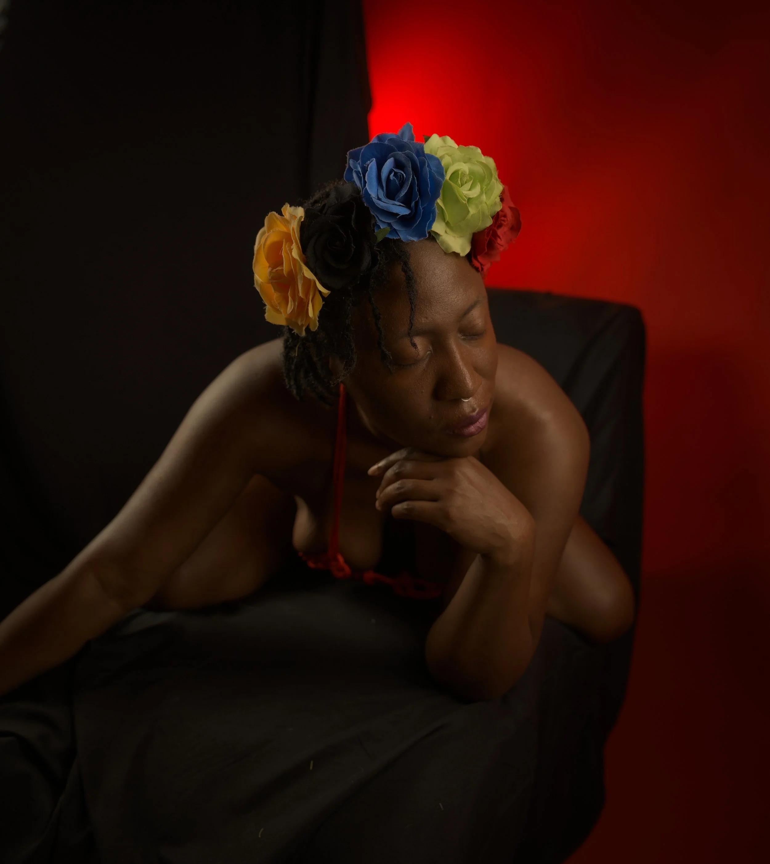 A woman with dark skin lying on her side with her eyes closed, wearing a colorful flower crown with yellow, blue, green, black, and red flowers, against a red and black background.