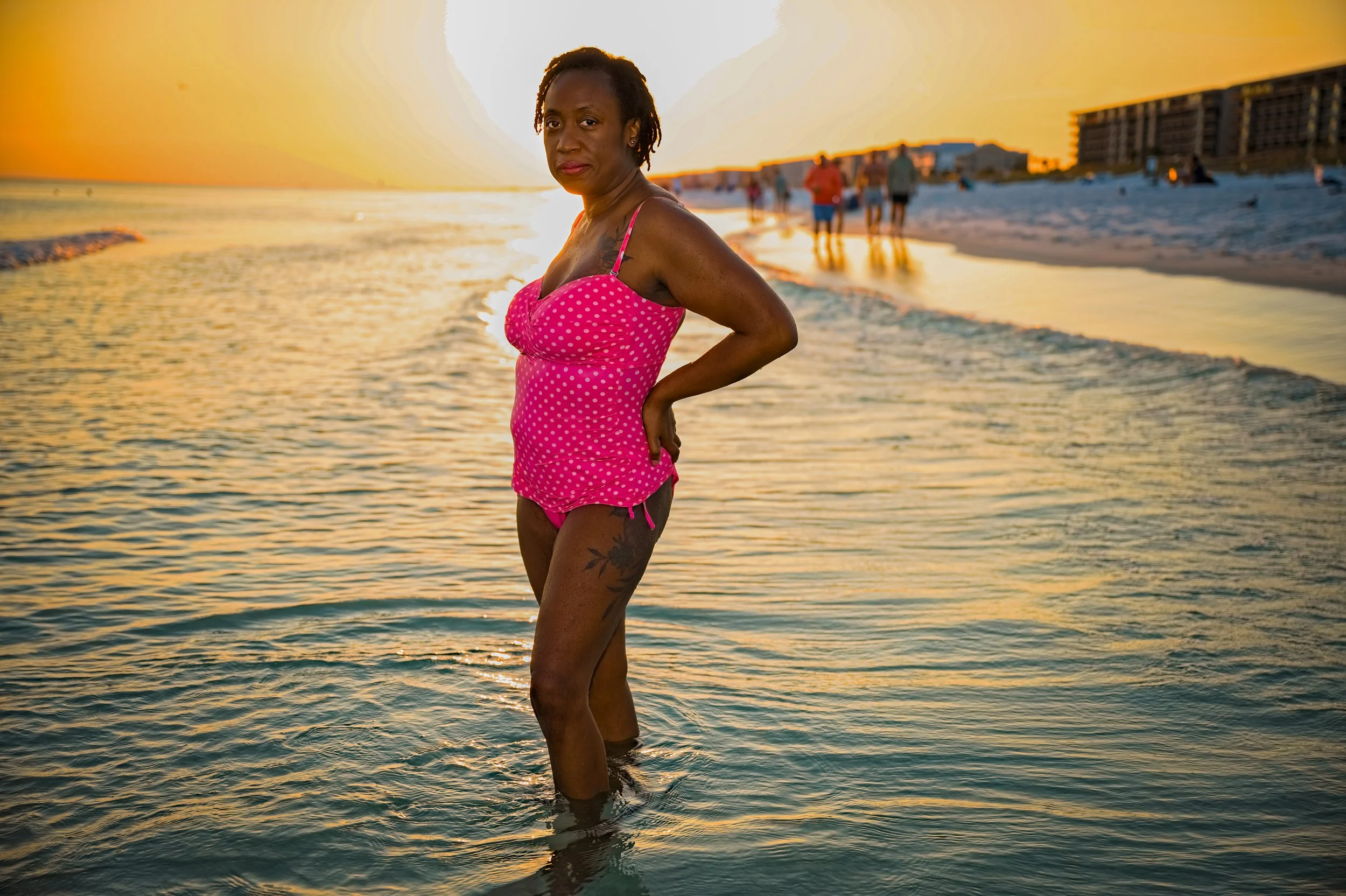 A woman standing in the shallow ocean water at sunset wearing a pink polka dot swimsuit, with a tattoo on her thigh, and people walking on the beach in the background.